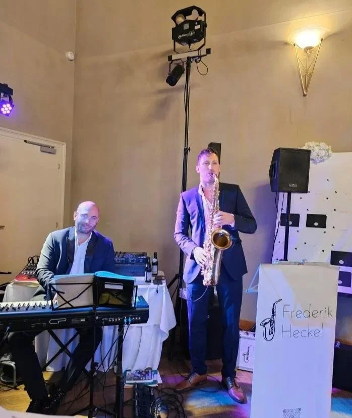 Zwei Musiker bei einer Live-Performance in einem Veranstaltungsraum, einer spielt Klavier und der andere Saxophon, mit Beschilderung "Frederik Heckel".