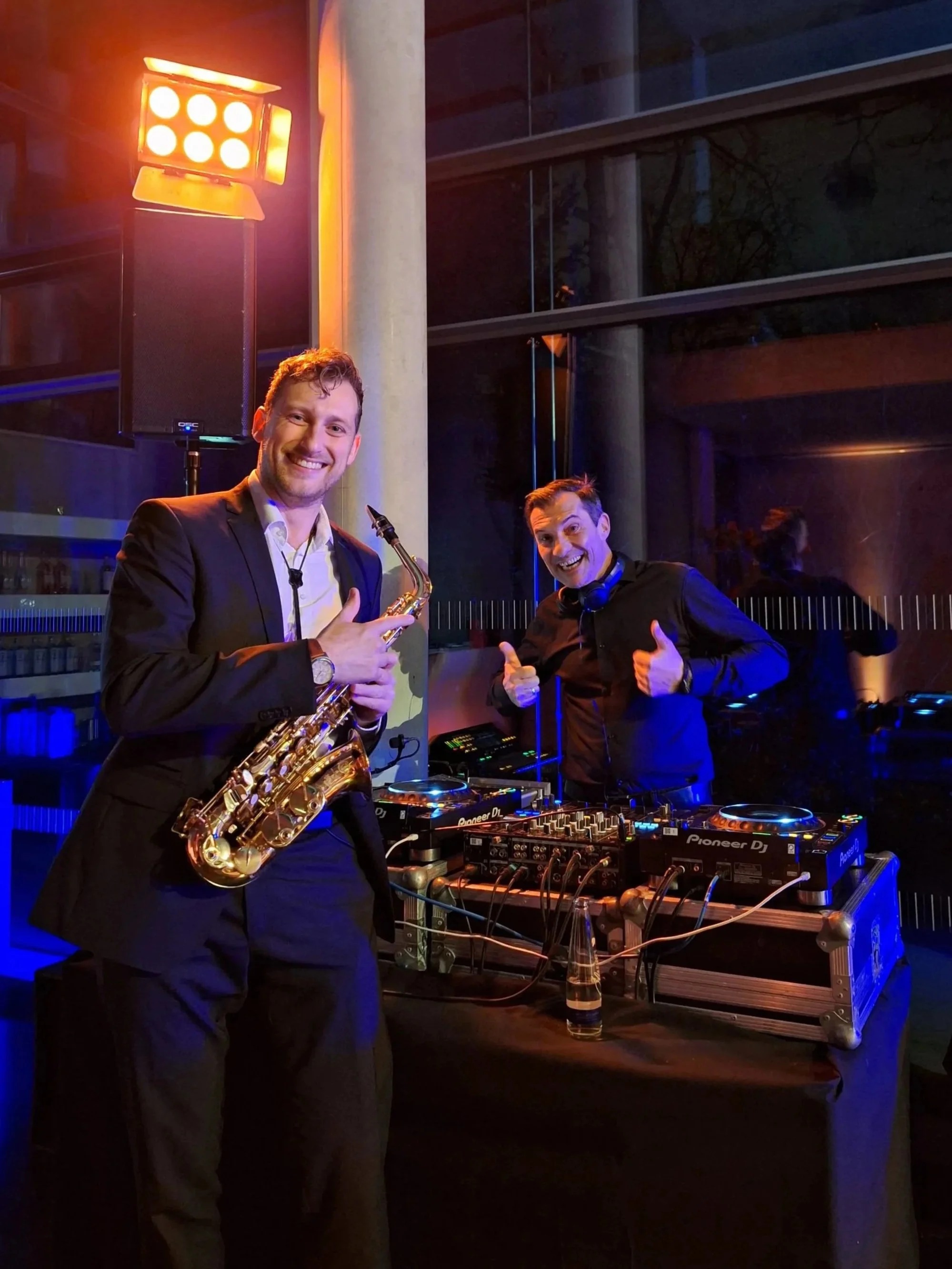 Zwei lachende Männer stehen an einem DJ-Pult, einer hält ein Saxophon, der andere zeigt Daumen hoch, im Hintergrund ein orangefarbener Bühnenstrahler.