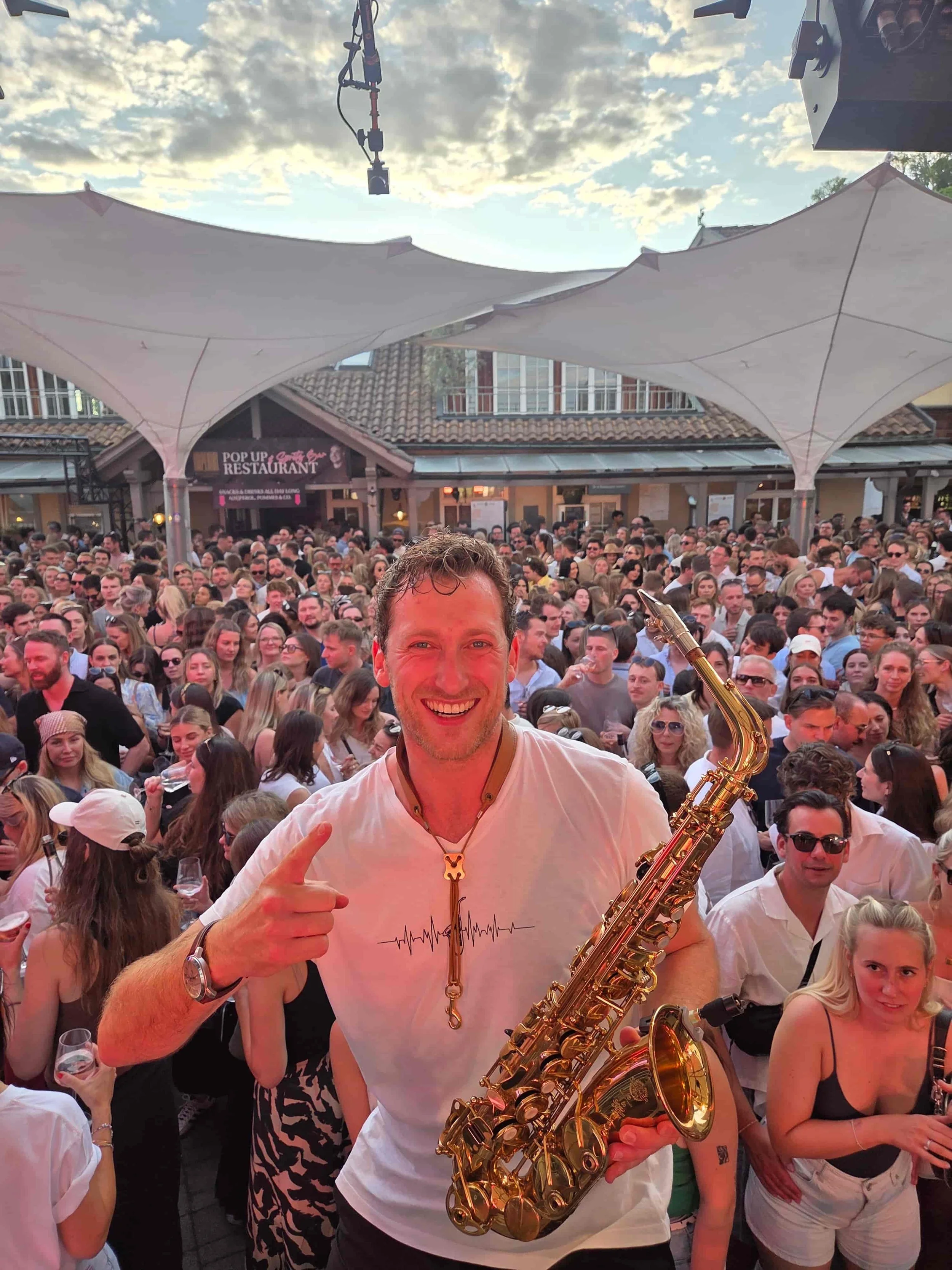 Ein lachender Mann hält ein Saxophon und zeigt auf sich selbst, während er in einer Menge Menschen bei einer Veranstaltung auf einer Terrasse steht. Im Hintergrund sind weiße Sonnensegel und ein Gebäude sichtbar.
