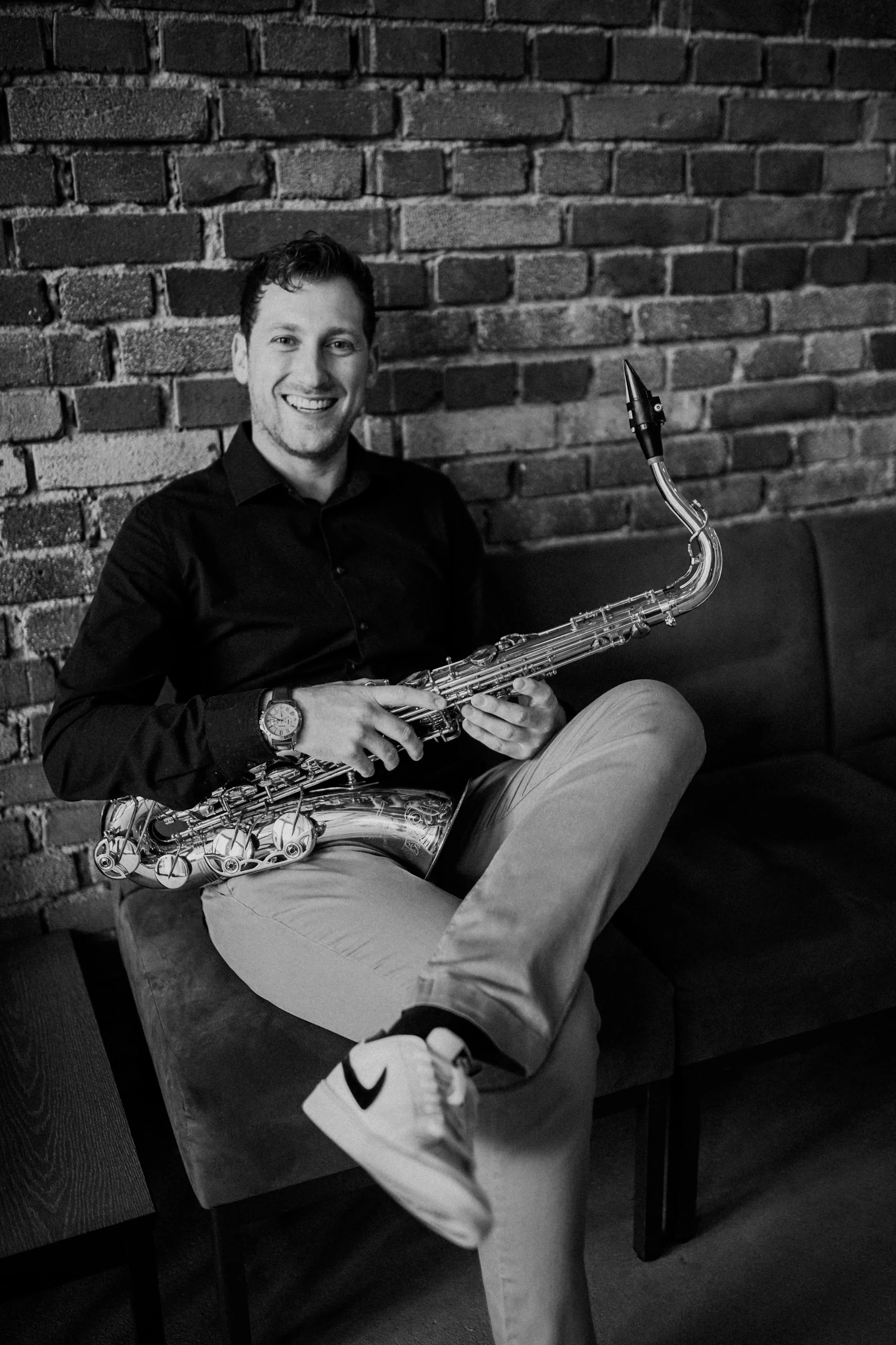Frederik Heckel in einem schwarzen Hemd und hellen Hosen sitzt auf einer Couch vor einer Ziegelsteinwand und hält eine Klarinette.