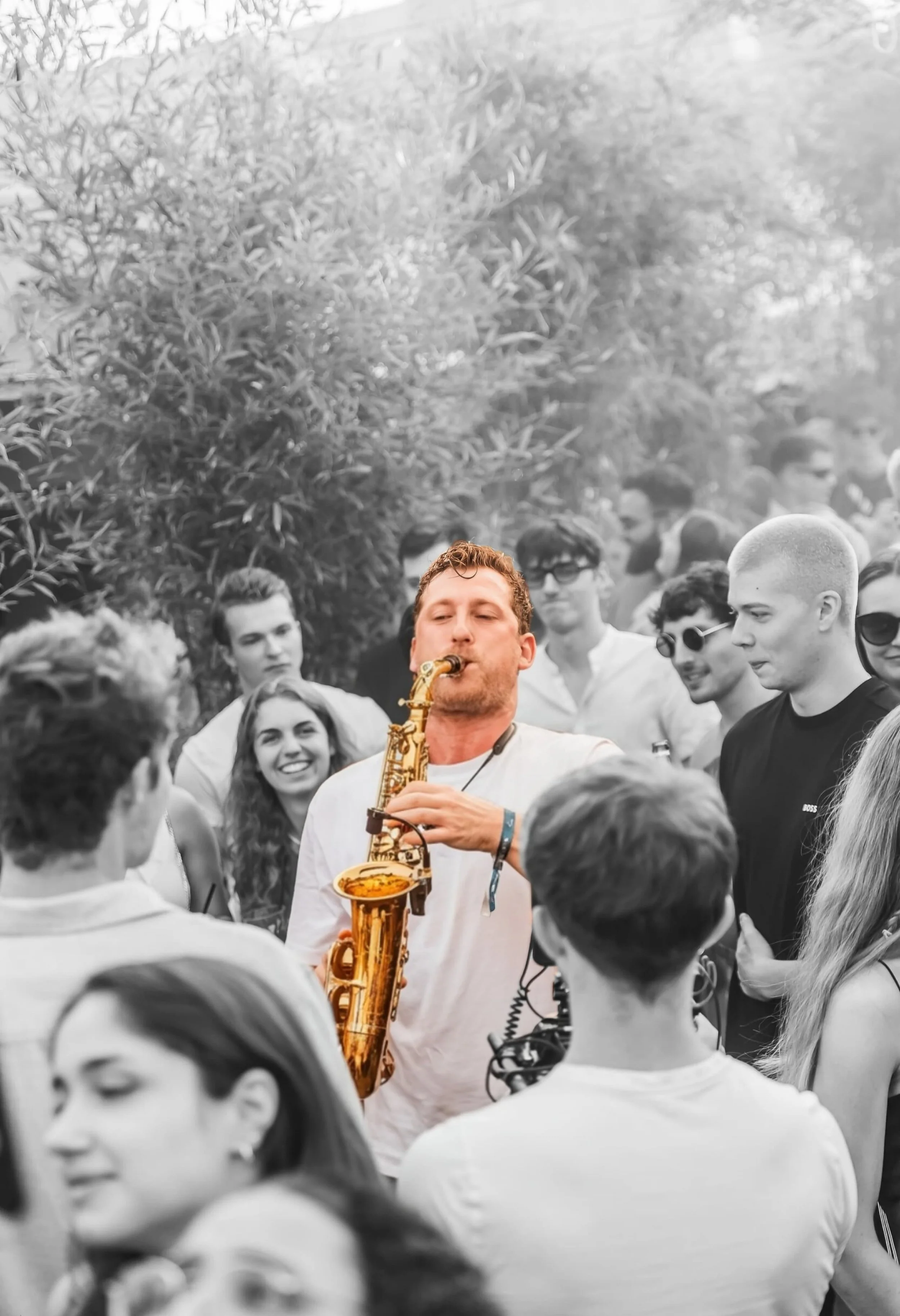 Frederik Heckel spielt Saxophon vor einer Gruppe lachender und bierender Menschen im Freien, mit Bäumen und Sonnenlicht im Hintergrund.