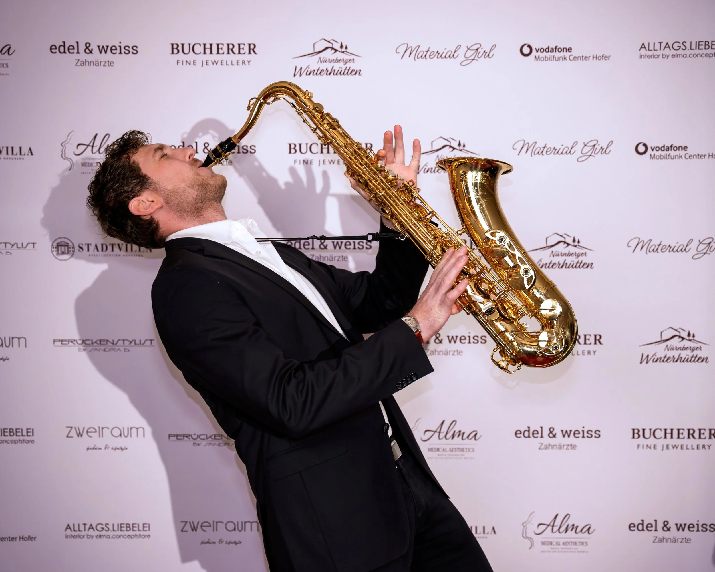 Frederik Heckel trägt einen schwarzen Anzug und spielt auf einem goldenen Saxophon bei einer Veranstaltung vor einer weißen Wand mit verschiedenen Logos.
