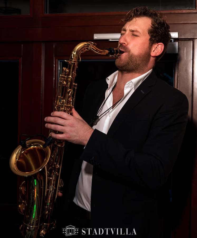 Frederik Heckel spielt auf einem goldenen Saxophon, trägt einen schwarzen Anzug und ein weißes Hemd, steht vor einer dunklen Holzwand.