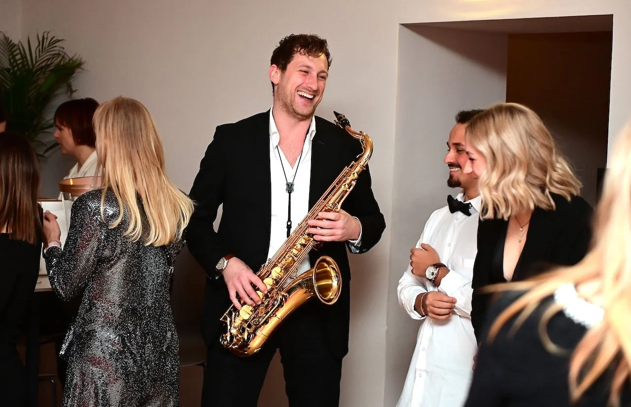 Menschen auf einer Party, Frederik Heckel mit Saxophon, alle lachen und haben Spaß.