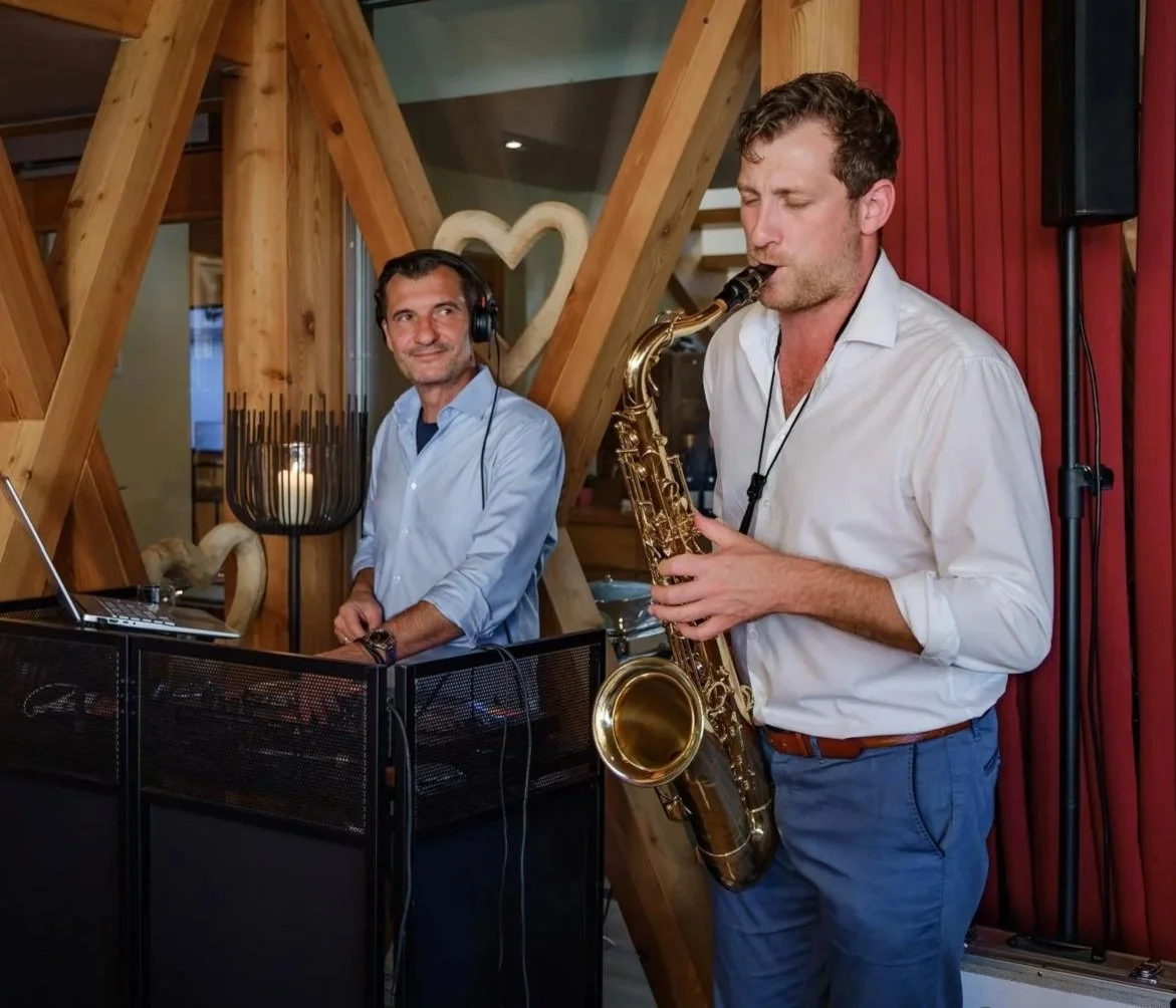 Frederik Heckel spielt Saxophon, ein anderer Mann steht neben einem DJ-Set mit Kopfhörern, in einem Raum mit Holzwanddekor und roten Vorhängen.