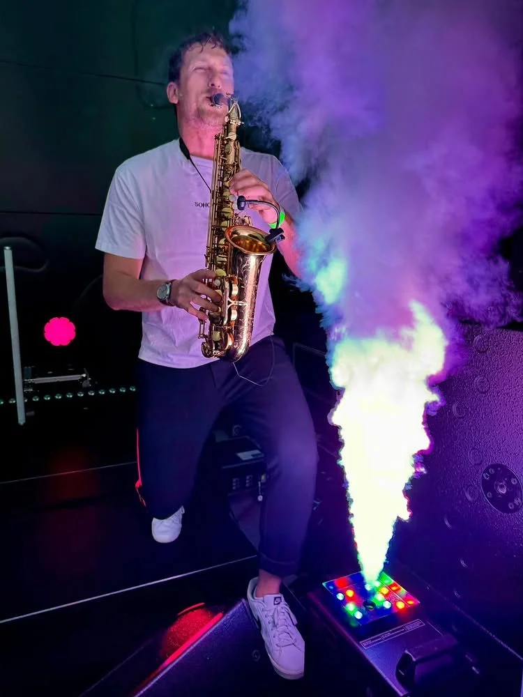 Frederik Heckel spielt Saxophon, während farbiges Rauch aus einer Maschine auf den Boden aufsteigt, in einer dunklen Umgebung.