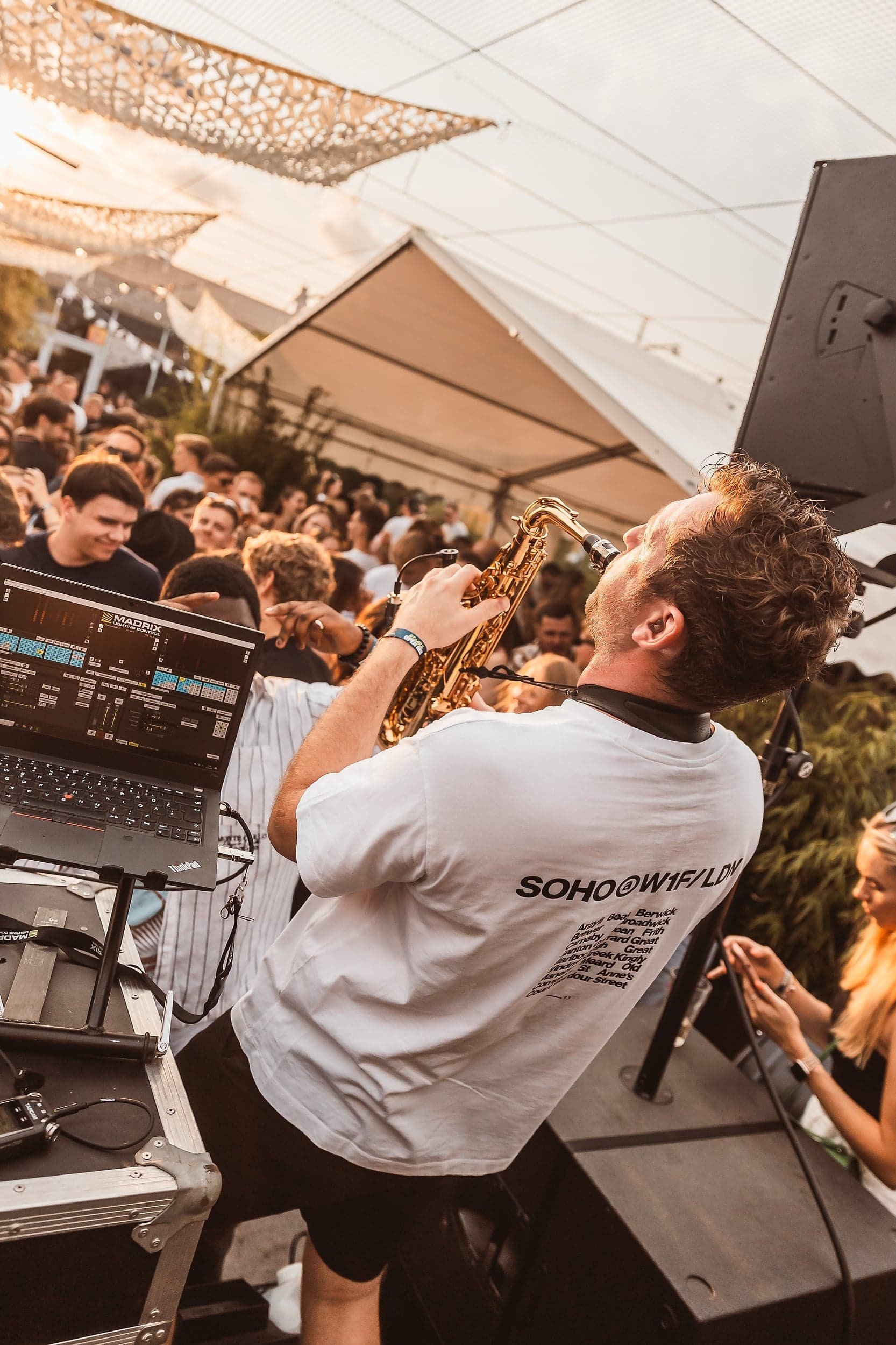 Ein DJ spielt auf einem Festival, während eine Band mit einem Saxophonist auftritt und das Publikum tanzt.