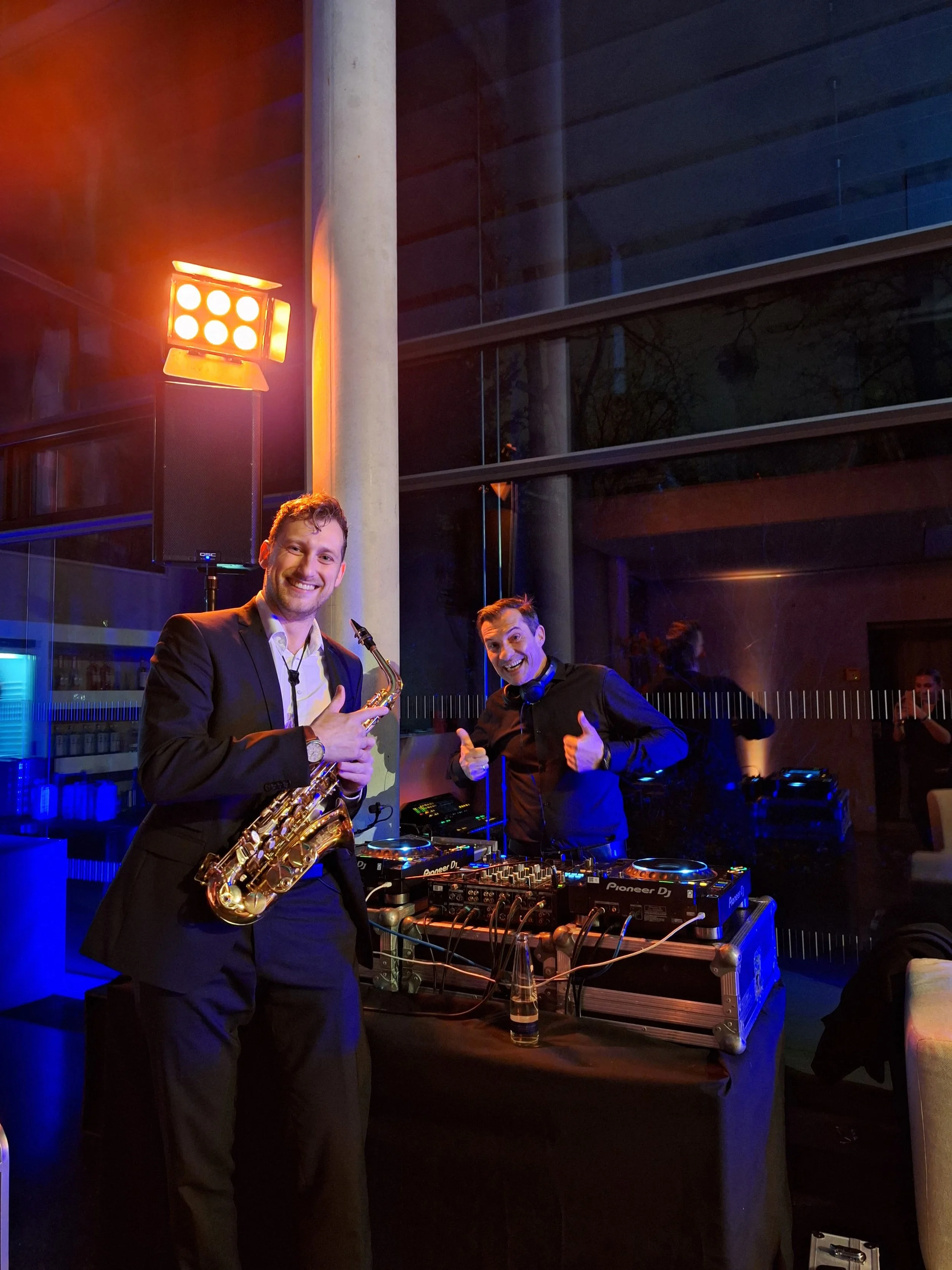 Zwei Männer bei einer Party. Einer hält ein Saxofon, während der andere an einem DJ-Setup arbeitet und beide lächeln. Es gibt bunte Lichter im Hintergrund.