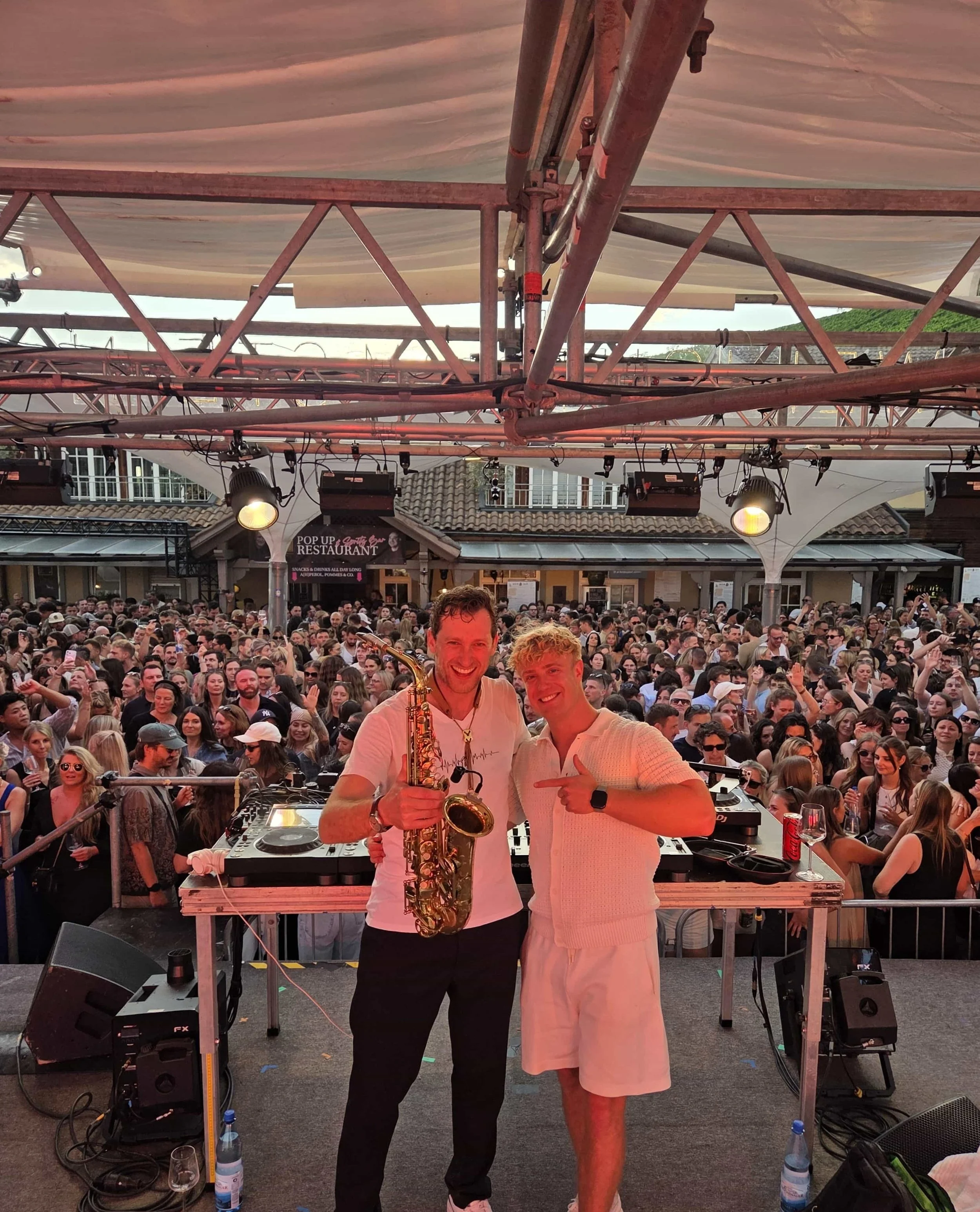 Zwei Männer, einer mit Saxophon, stehen an einem DJ-Pult auf einer Bühne vor einer großen Menschenmenge bei einem Open-Air-Festival. Das Bühnenbild ist mit Licht- und Tontechnik ausgestattet, im Hintergrund sind Dach und Gebäude sichtbar.