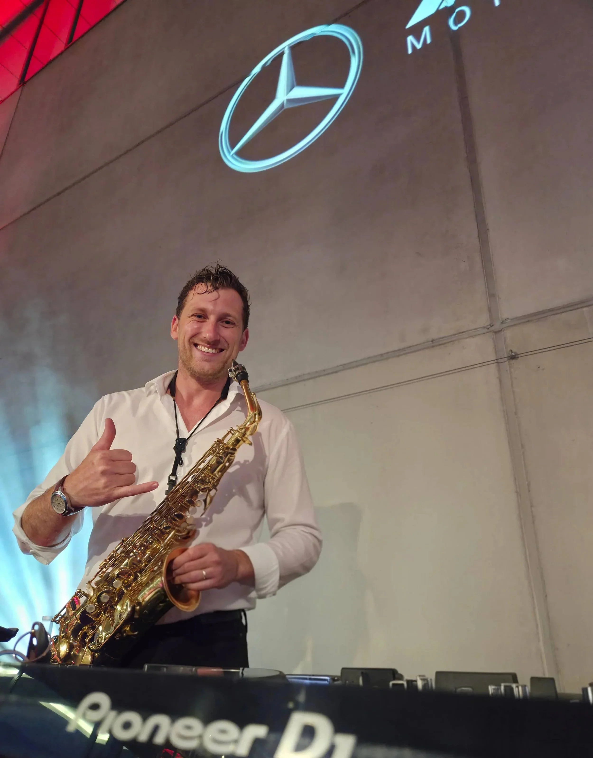 Frederik Heckel mit Saxophon, der eine Hand zum Daumen hoch hebt, steht vor einer DJ-Station bei einer Veranstaltung, im Hintergrund eine Projektion des Mercedes-Benz-Logos an eine Wand.