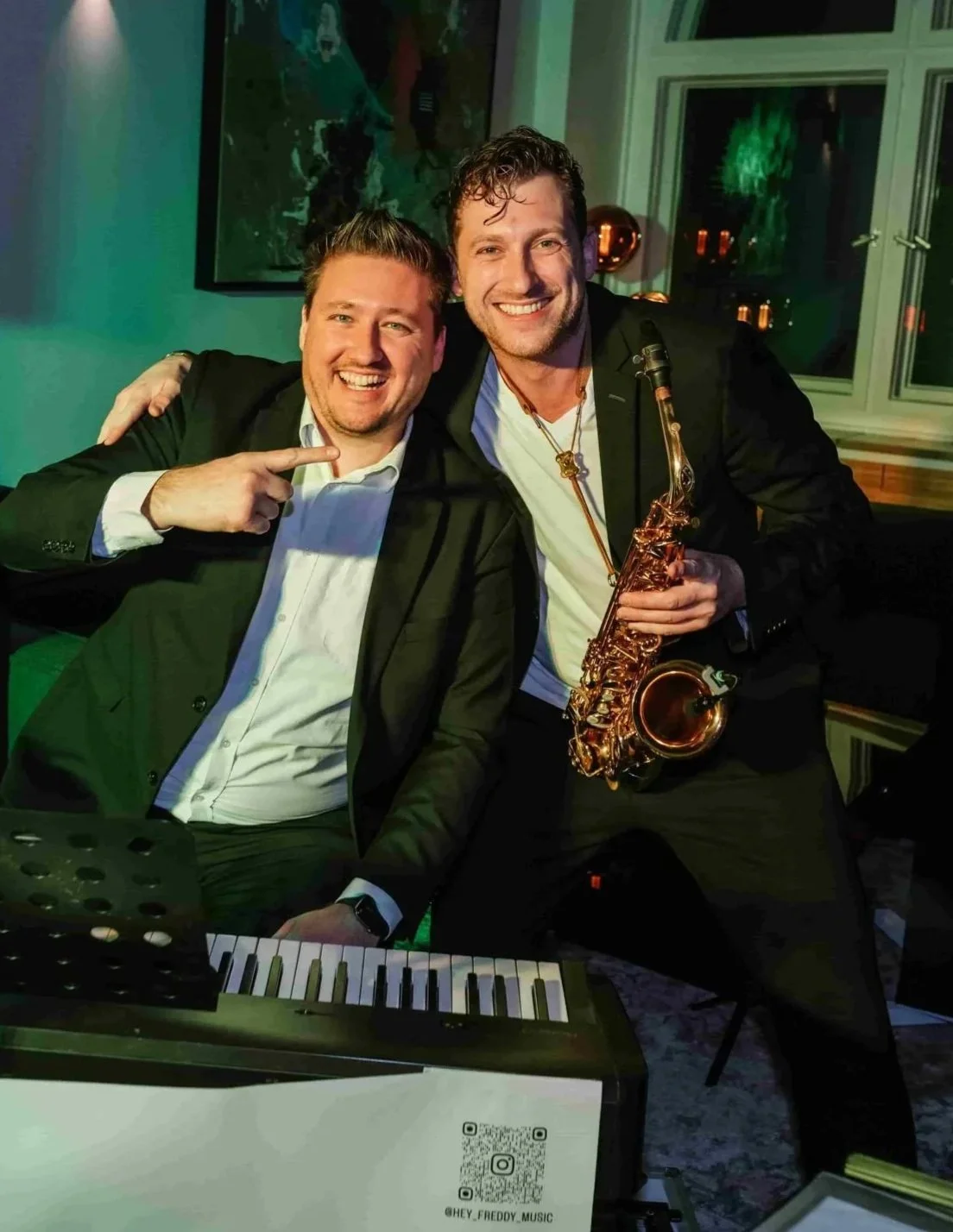 Zwei Männer in Anzügen, lachen und posieren mit einem Saxophon und einem Keyboard in einer festlichen Umgebung.