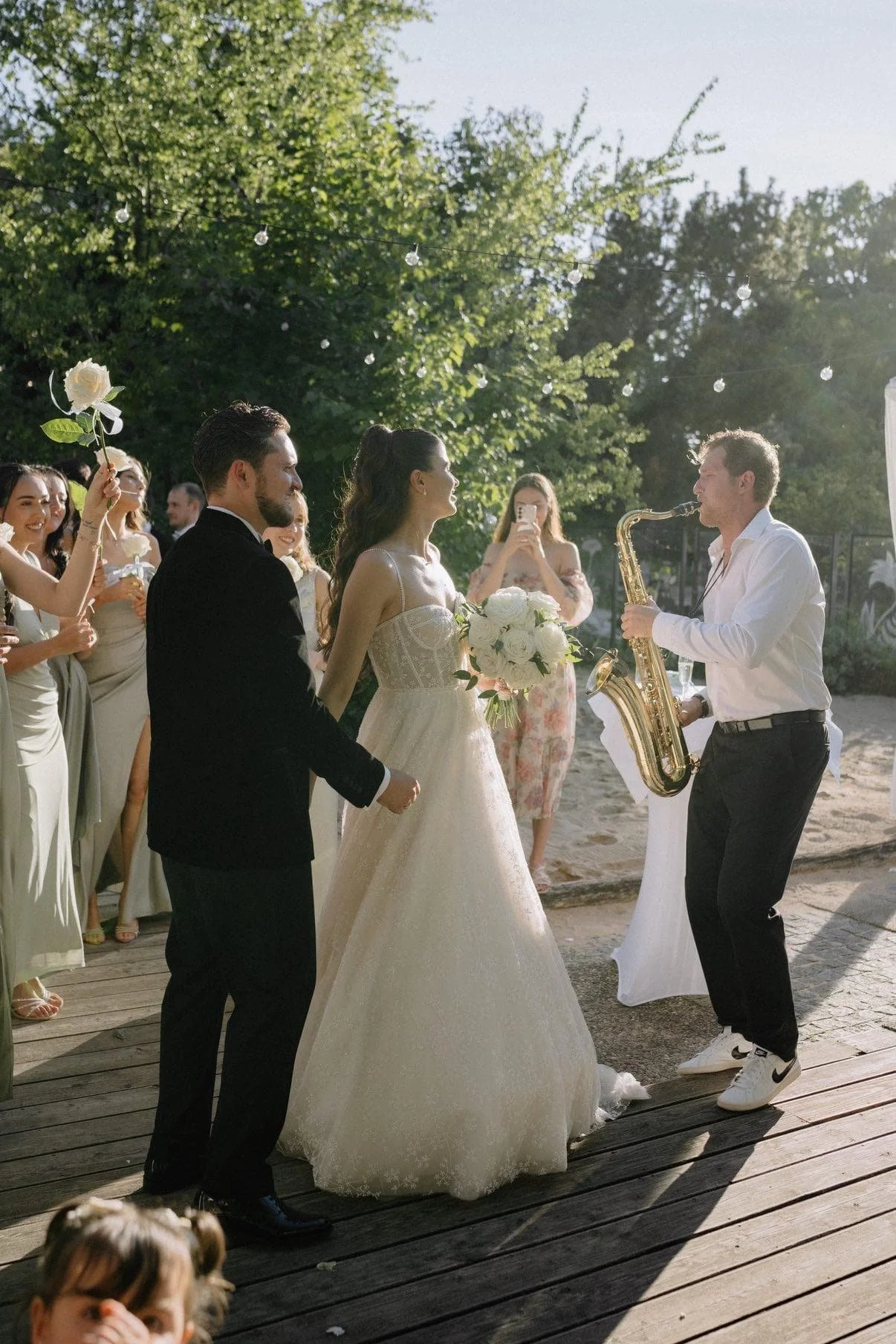 Hochzeit im Freien mit Braut im weißen Kleid, Bräutigam in schwarzem Anzug, Frederik Heckel mit Saxophon, Gäste mit Blumen und Fotos im Sonnenlicht.