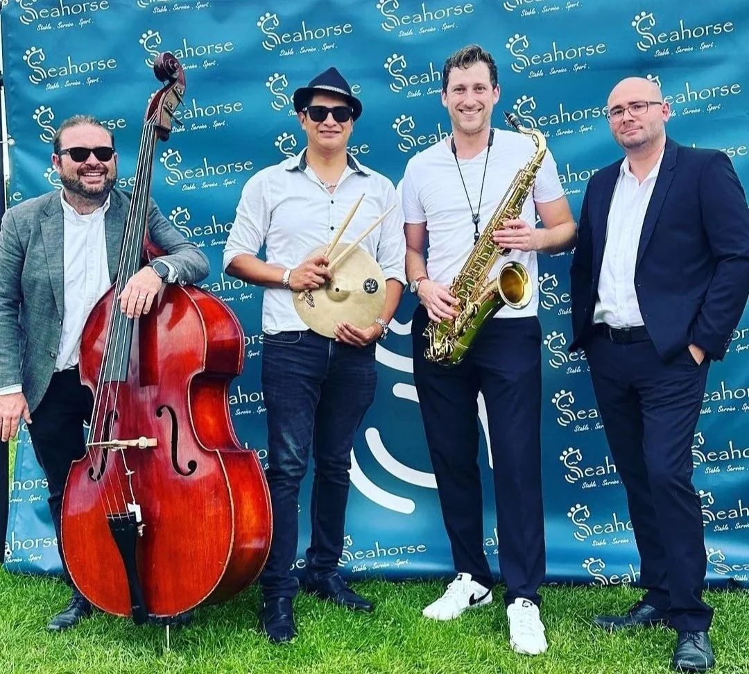 Vier Männer, Band aus Nürnberg, stehen vor einem blauen Hintergrund mit dem Logo 'Seahorse'. Jeder hält ein Musikinstrument: Kontrabass, Becken, Saxophon, und einer hat keine sichtbare Waffe. Sie tragen formelle Kleidung, und alle lachen.