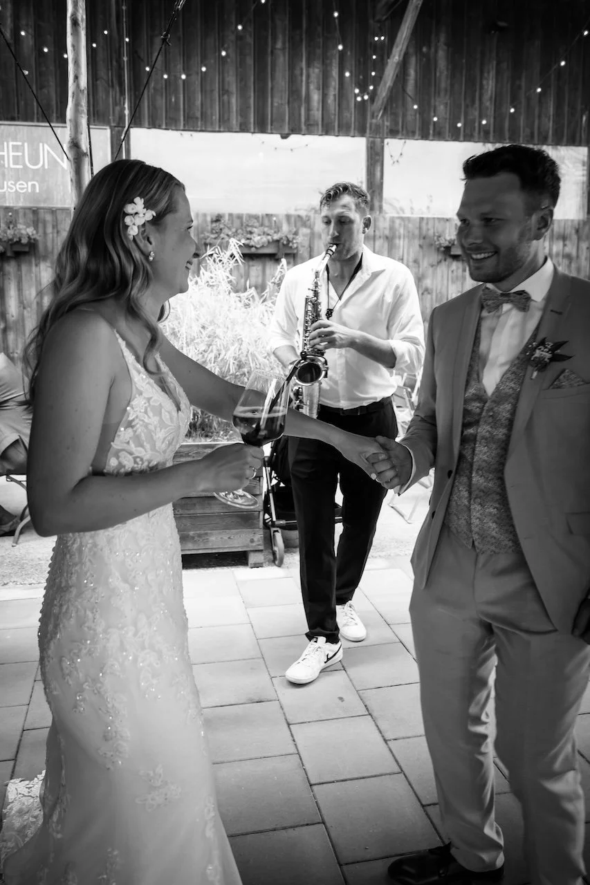 Ein Brautpaar bei einer Hochzeit, das sich die Hand gibt, während Frederik Heckel im Hintergrund auf einem Saxophon spielt.