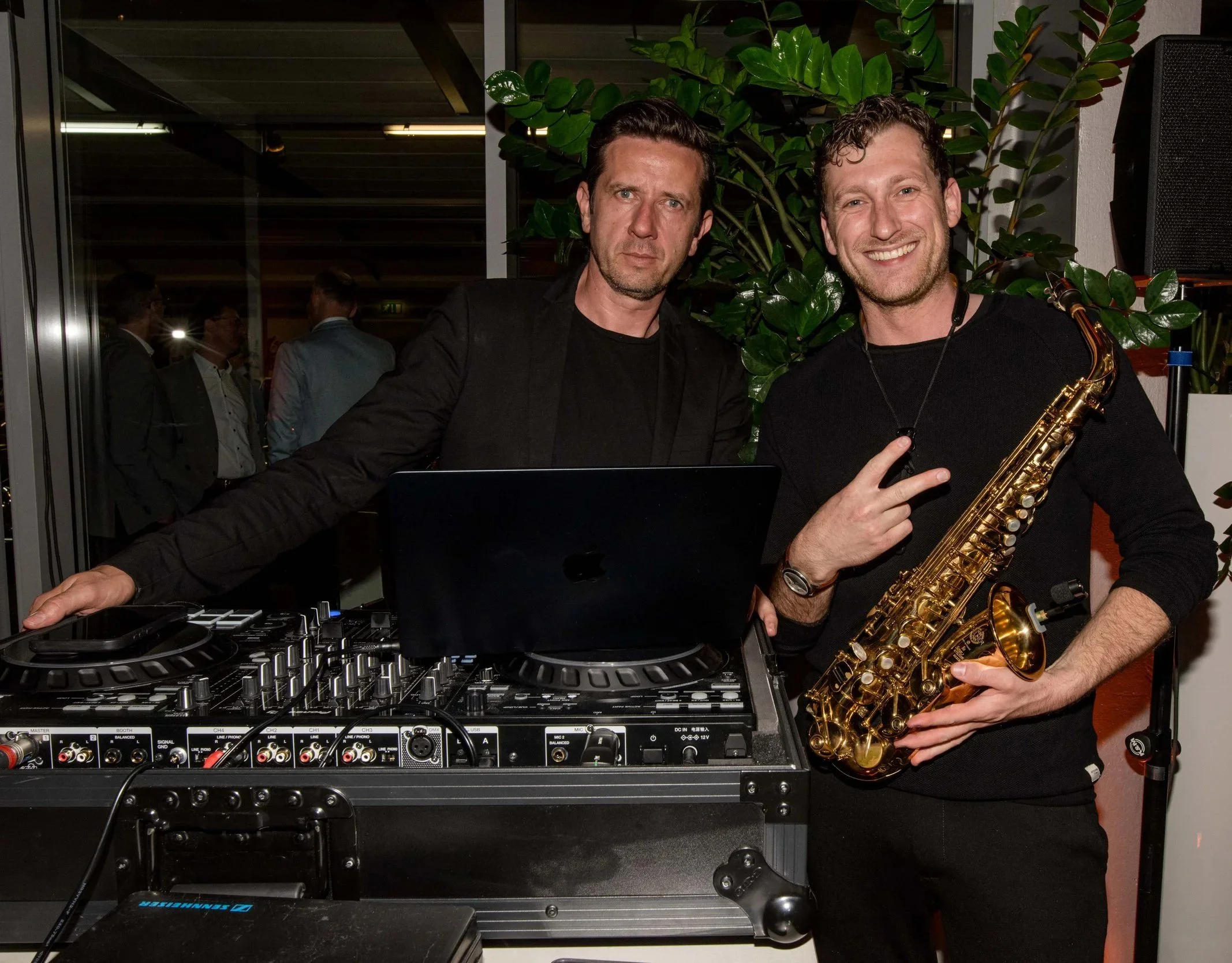 Zwei Männer, einer mit DJ-Equipment und der andere mit Saxophon, feiern bei einer Veranstaltung.