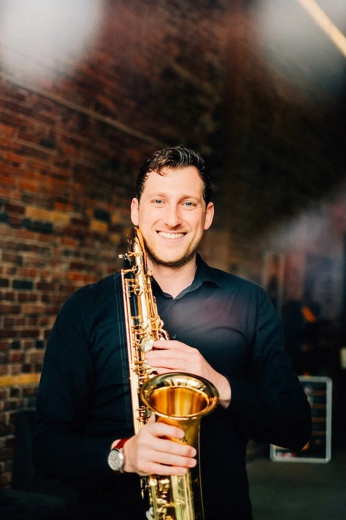 Frederik Heckel hält ein Saxophon und lächelt in einem Raum mit Backsteinwand im Hintergrund.