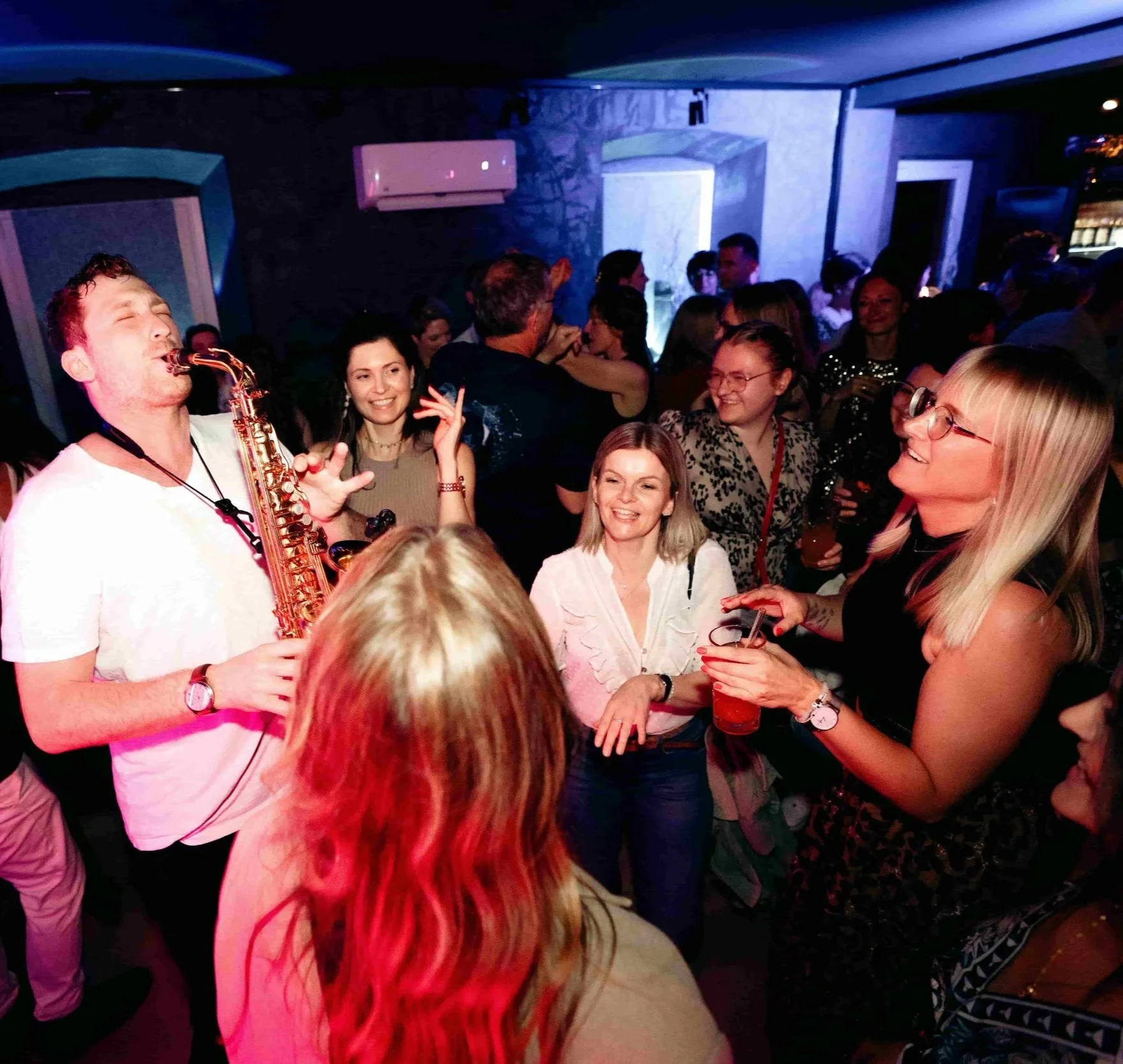 Menschen feiern in einer Bar, Frederik Heckel spielt Saxophon, andere lachen und tanzen, es herrscht eine lebhafte Atmosphäre.
