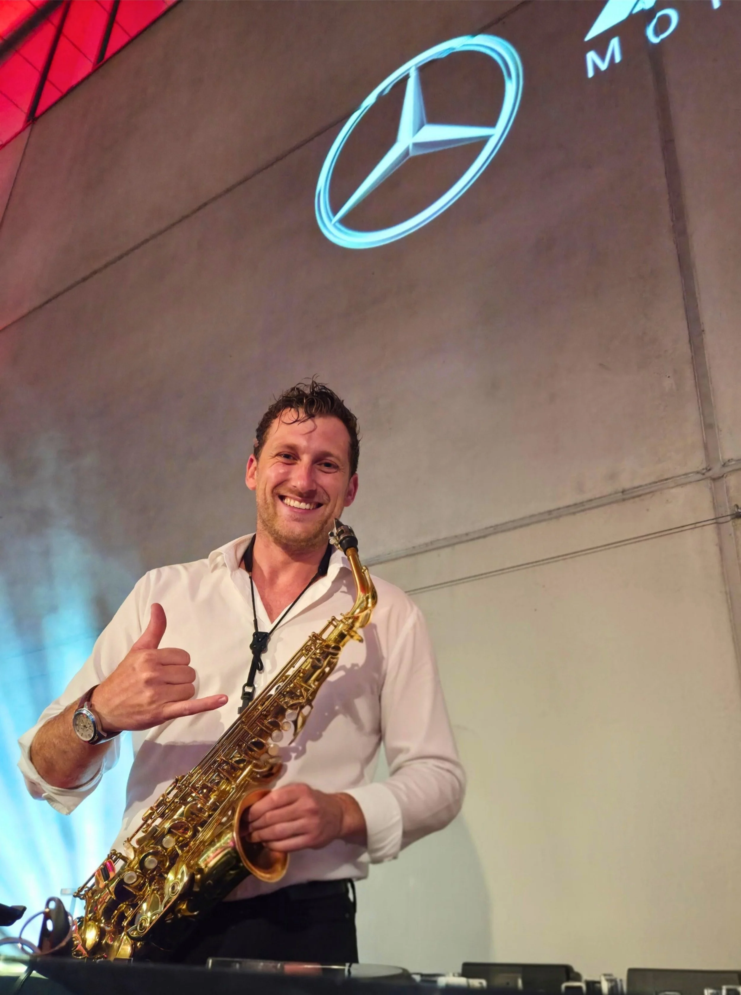 Ein lachender Mann mit einem Saxophon in der Hand, macht ein OK-Handzeichen und posiert vor einer grauen Wand mit einem leuchtenden Mercedes-Benz-Logo an der Decke.