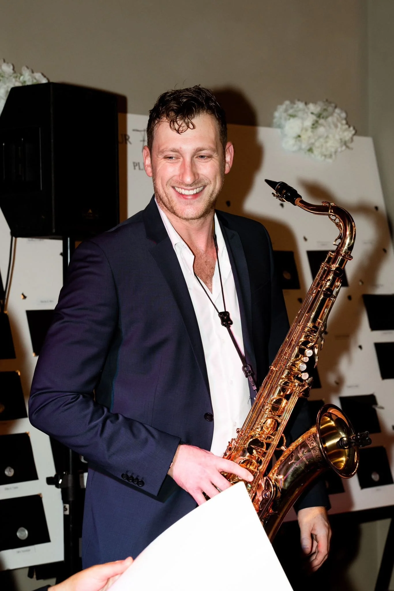 Frederik Heckel mit Saxophon bei einer Feier, lachend, in formeller Kleidung, im Hintergrund Notenständer.