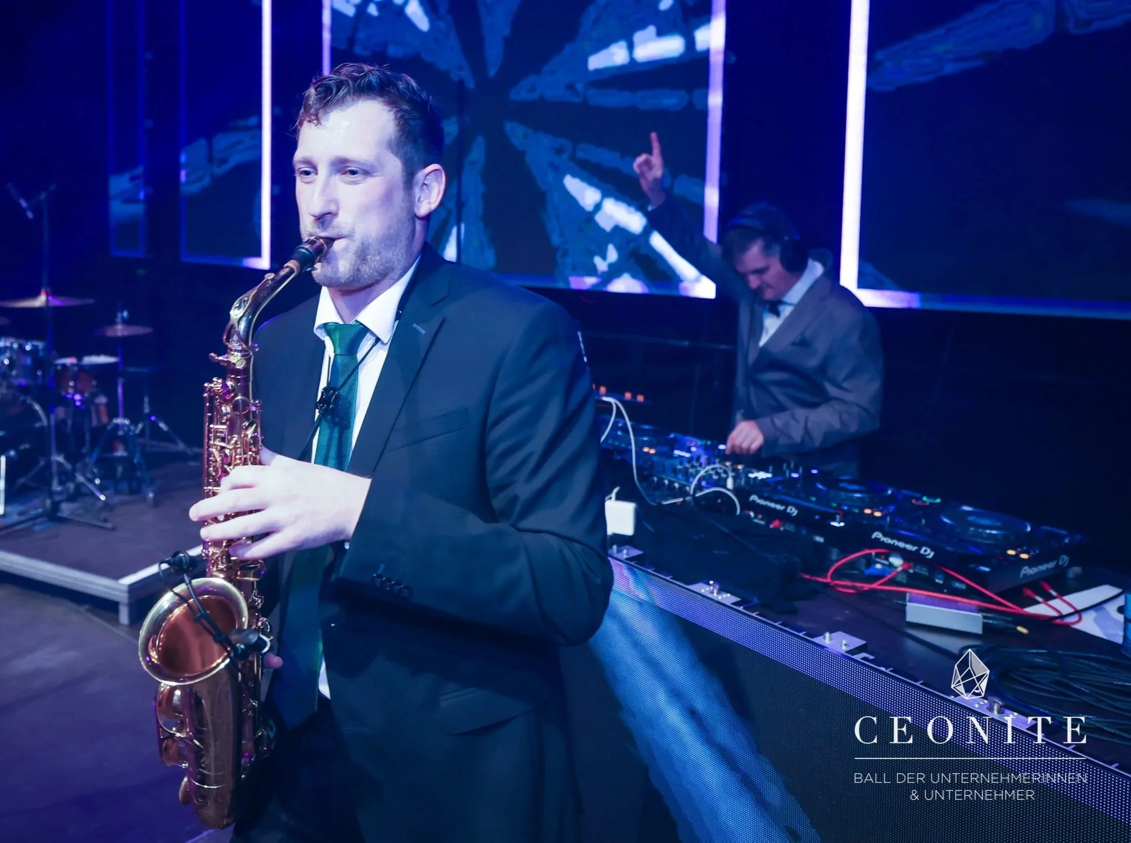 Frederik Heckel beim Auftritt, Mann spielt Saxophon, im Hintergrund DJ an Turntables, dunkle Bühne mit buntem Licht, CEONITE Logo im unteren rechten Eck, Text in Deutsch.