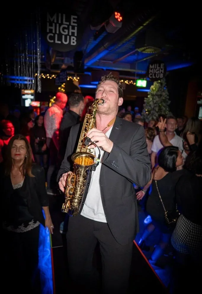 Frederik Heckel spielt Saxophon in einer Party-Atmosphäre mit Menschen im Hintergrund, bunte Lichter und Dekorationen, im Mile High Club