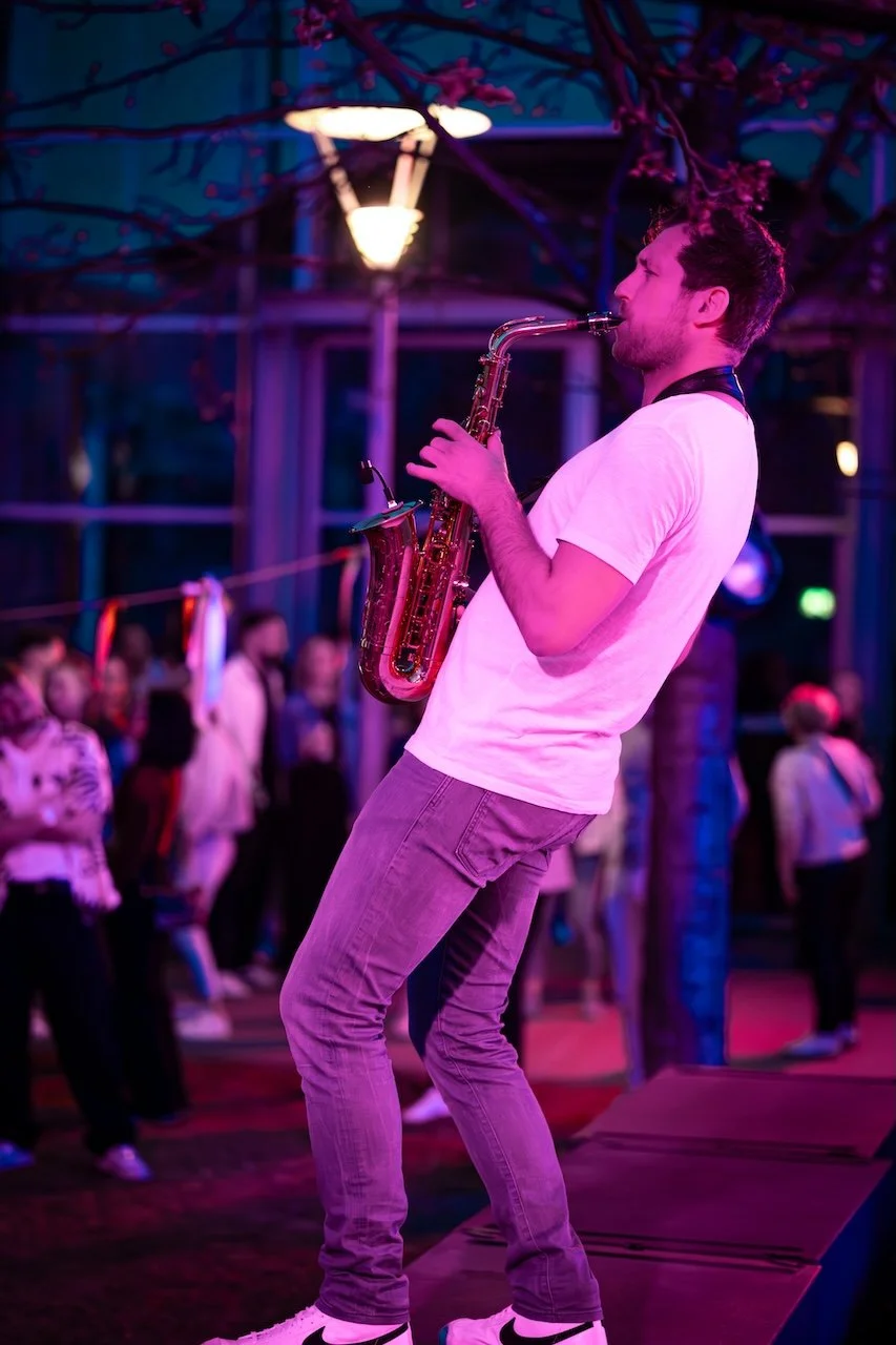 Frederik Heckel spielt Saxophon auf einer kleinen Bühne, während im Hintergrund eine Gruppe von Leuten im Dunkeln beobachtet.