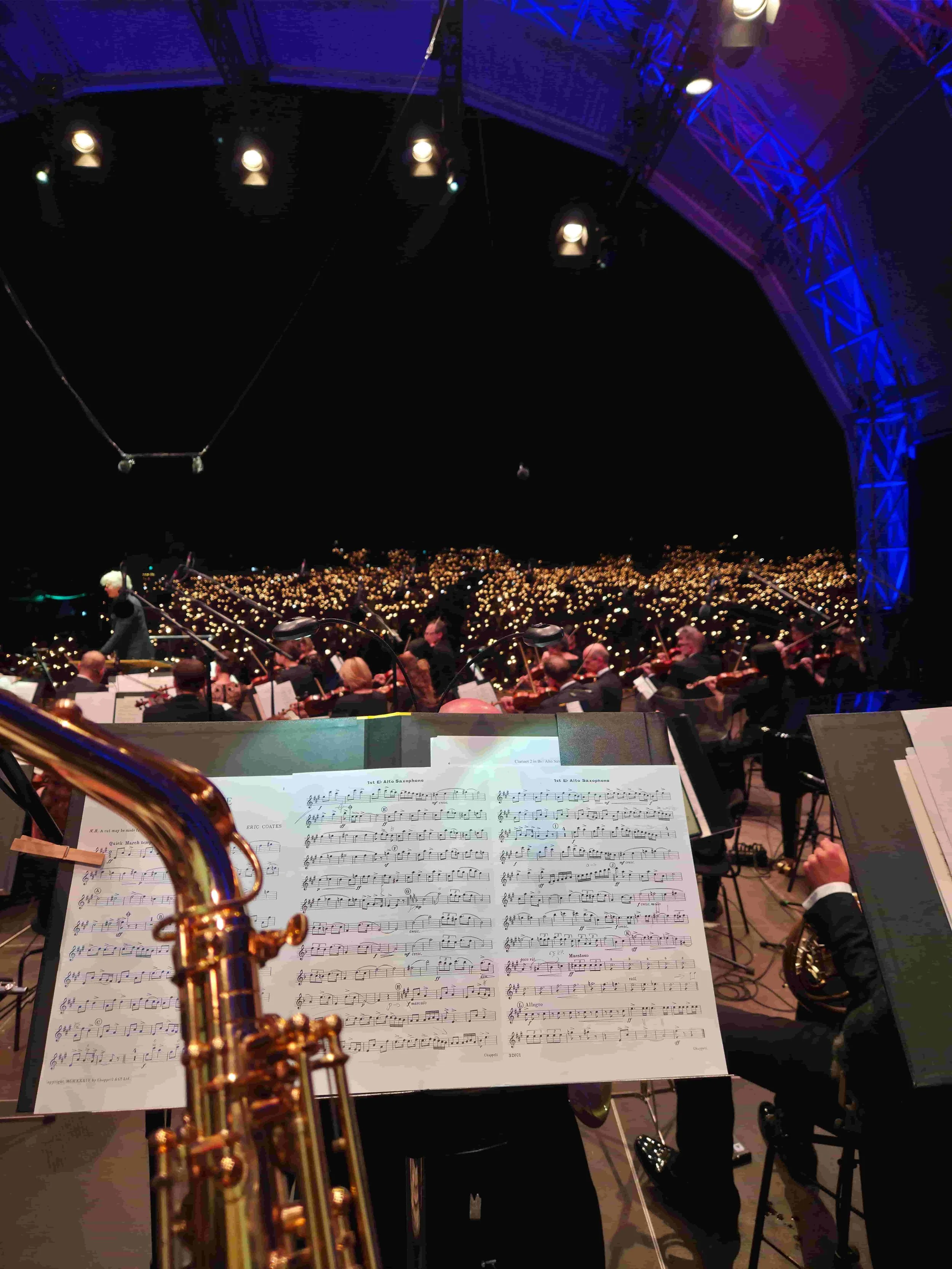 Orchester bei einem Konzert, aufgenommen aus Blickrichtung des Dirigenten, mit Notenständern, Musikern und einem Publikum mit Lichtern im Hintergrund.
