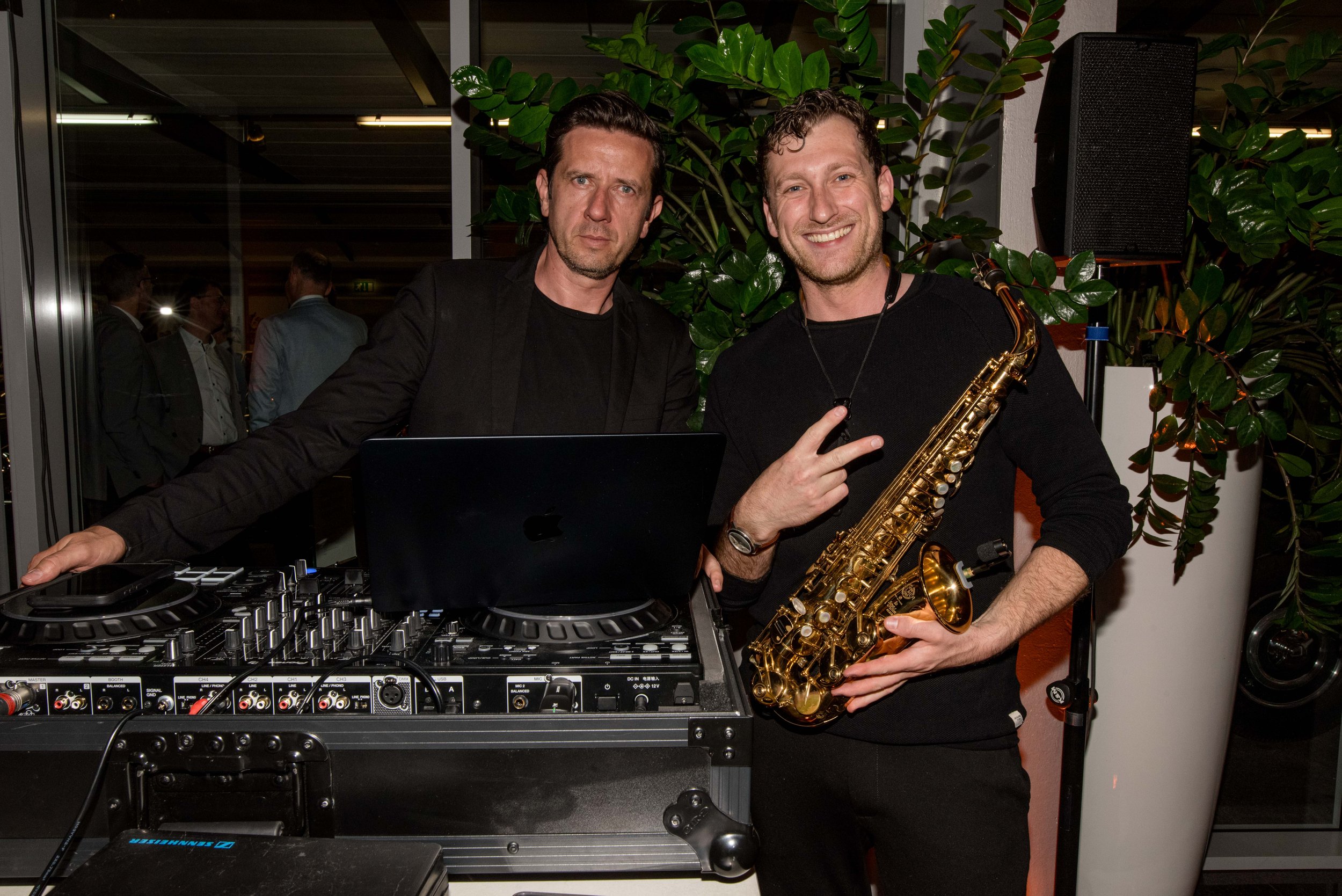 Zwei Männer, einer mit einem DJ-Controller und einem Laptop, der andere mit einem Saxophon, stehen nebeneinander bei einer Feier, umgeben von grünen Pflanzen, im Hintergrund sind andere Gäste zu sehen.