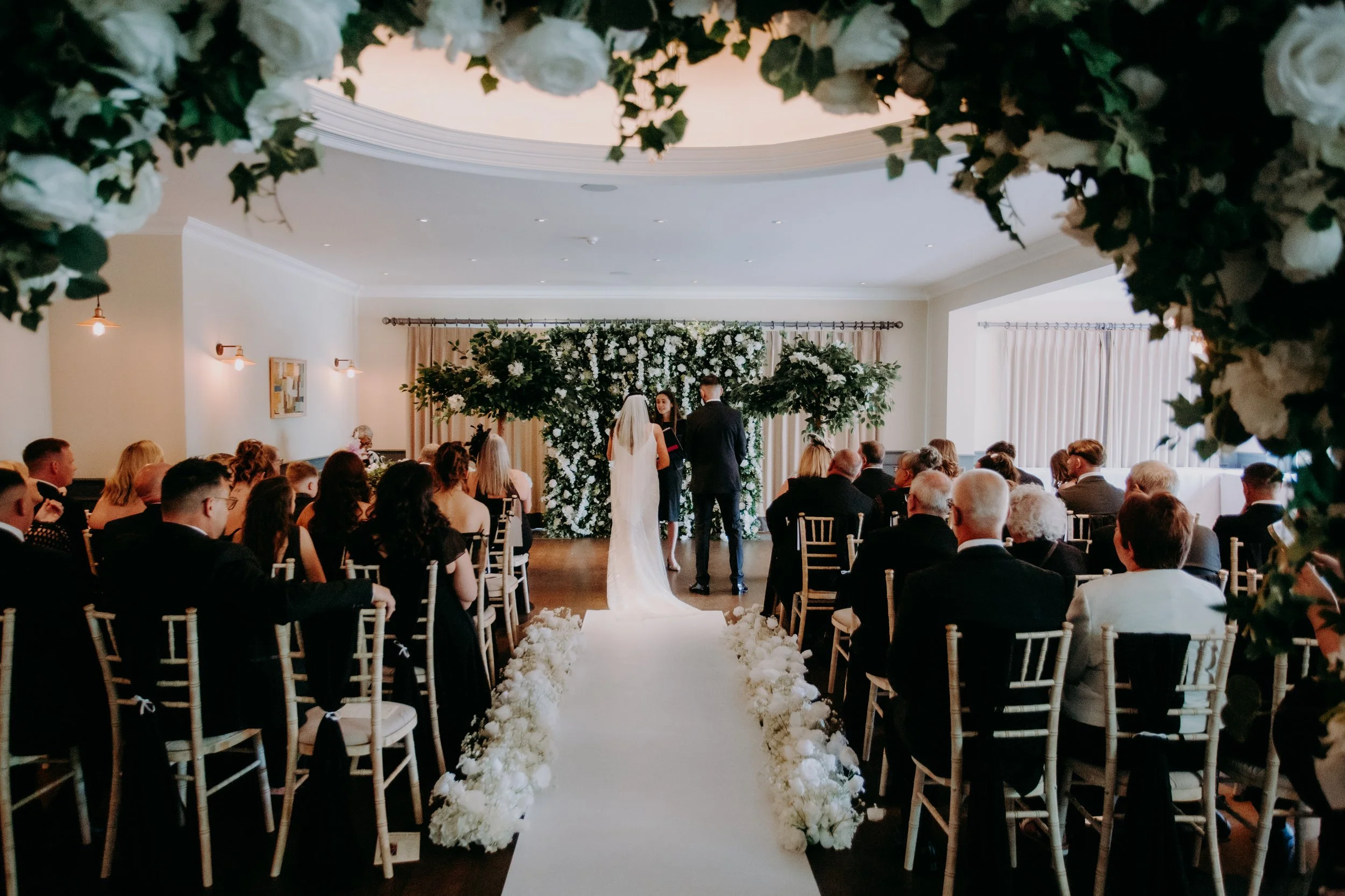 Wedding ceremony with floral arch and guests seated in an elegant indoor venue.