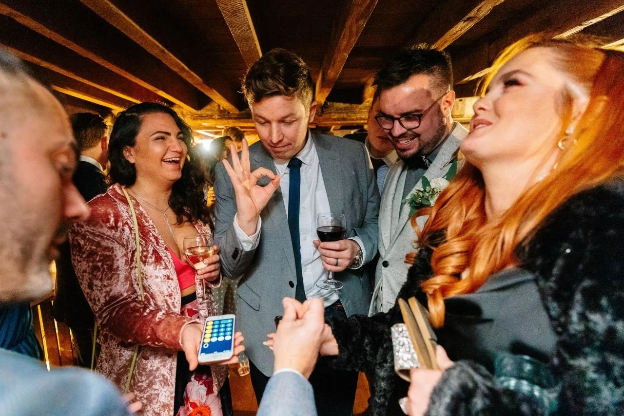 Wedding guests laughing and reacting during close-up magic performance in a rustic reception space.