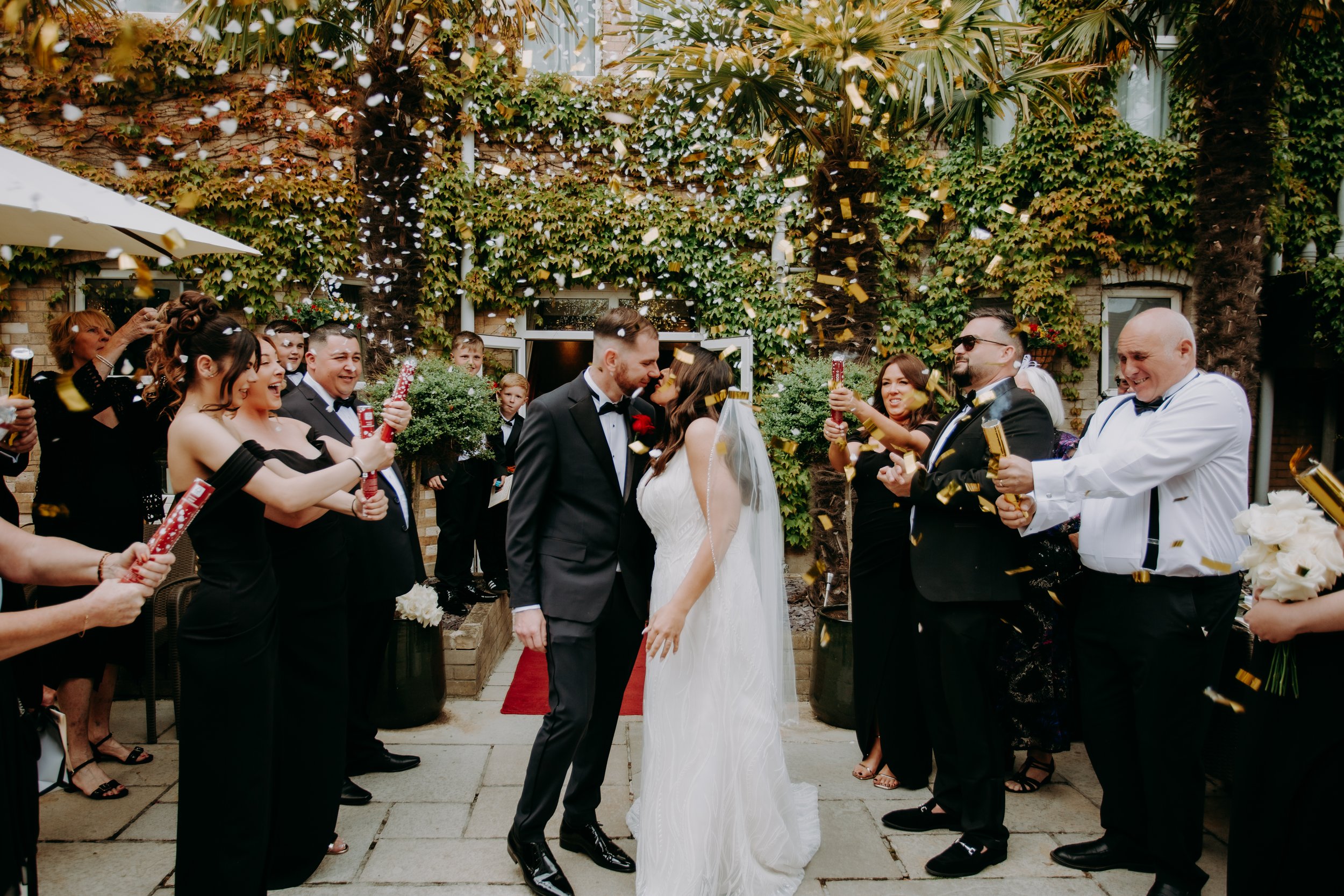 Bride and groom kissing during confetti exit surrounded by wedding guests.