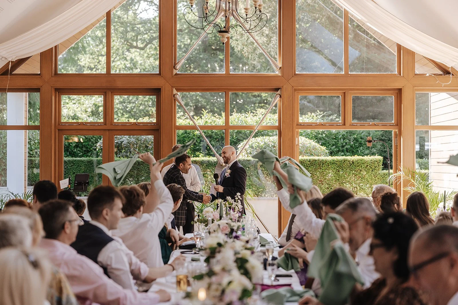 Groom assisting magician during a cabaret magic finale as wedding guests raise napkins and laugh inside a bright glass reception space.