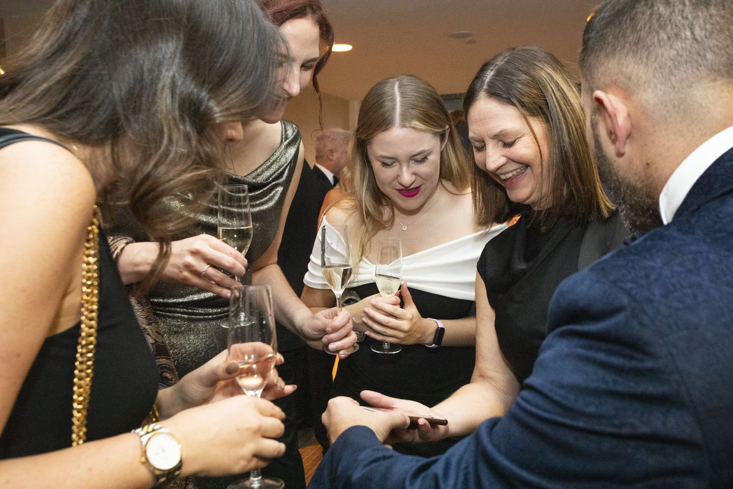A magician in a navy velvet blazer performs a close-up card trick for a group of guests at a corporate black-tie event, drawing laughter and amazement from the audience.
