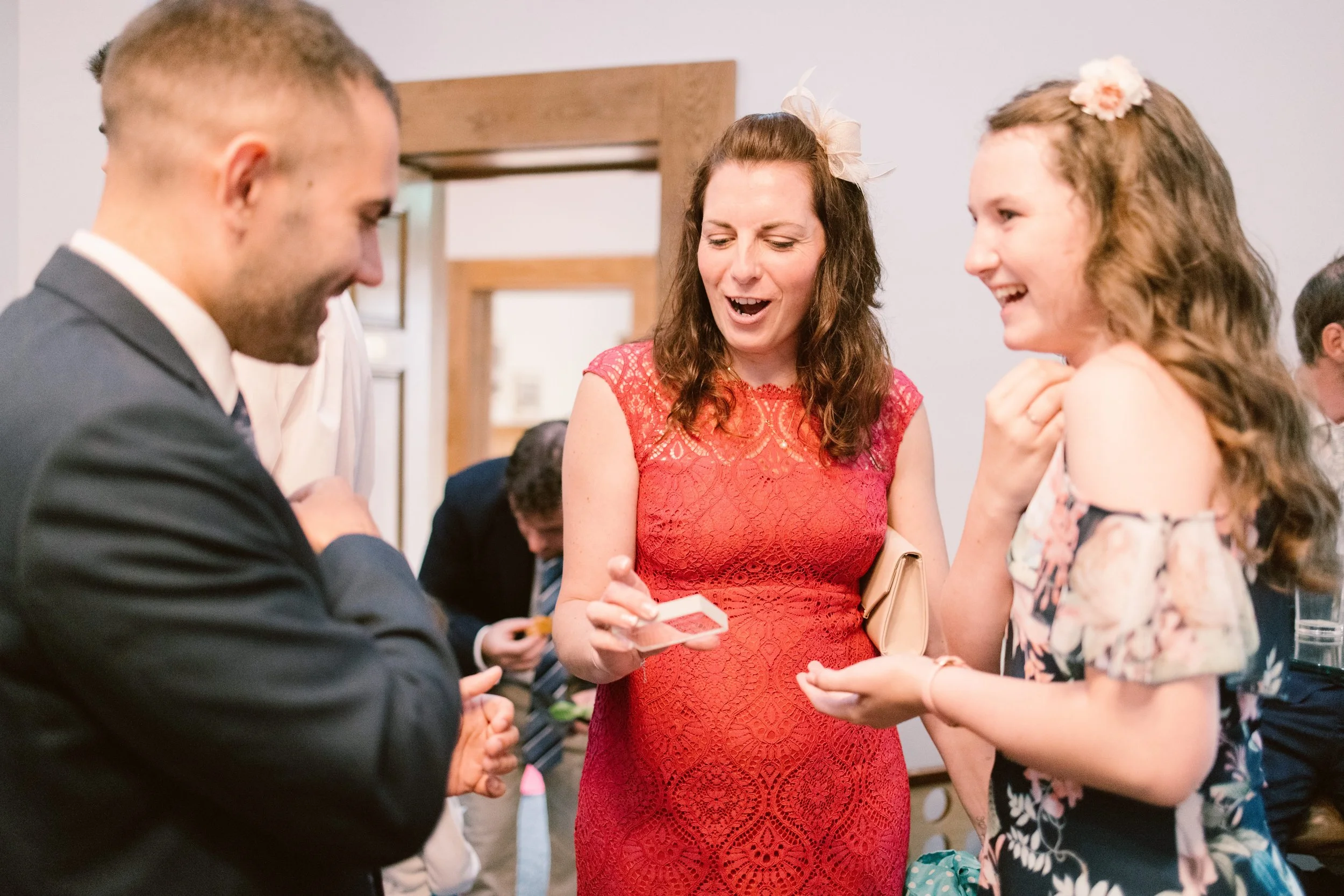 A wedding guest in a red lace dress reacts with open-mouthed surprise while holding a playing card during a close-up magic trick at a wedding reception