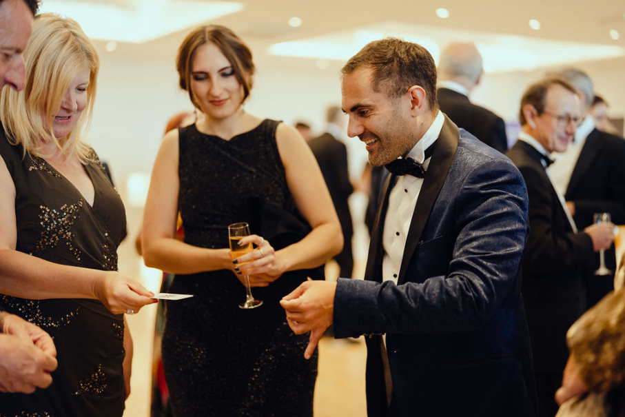 A magician in a navy velvet blazer performs a close-up card trick for a group of guests at a corporate black-tie event, drawing laughter and amazement from the audience.