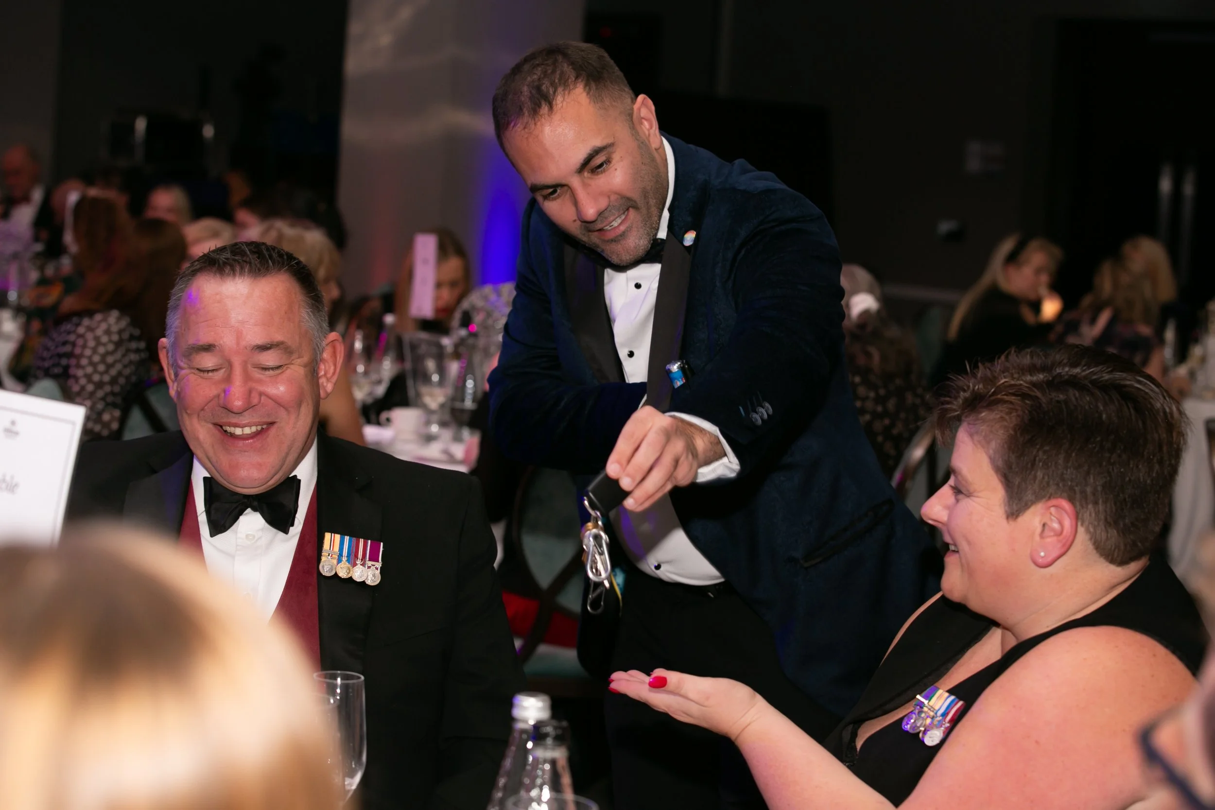 A magician in a navy tuxedo performs close-up magic for guests seated at a table during a black-tie corporate awards dinner, drawing laughter and amazement from the audience.