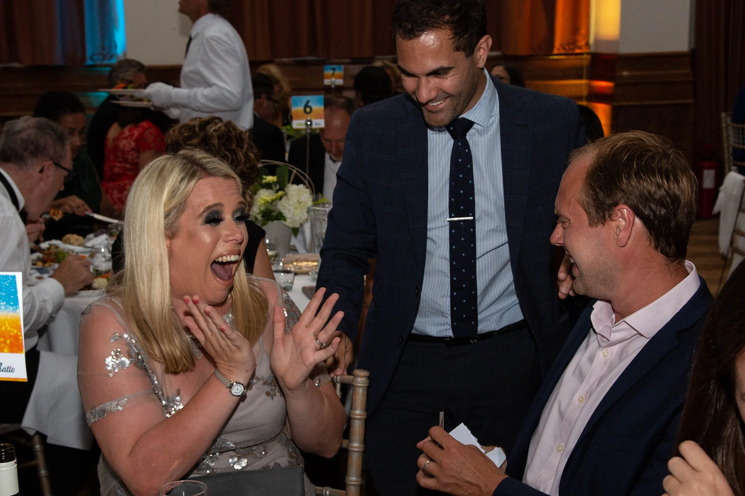 A magician in a suit performs close-up magic for guests seated at a table during a black-tie awards dinner, drawing surprised and delighted reactions from the audience.