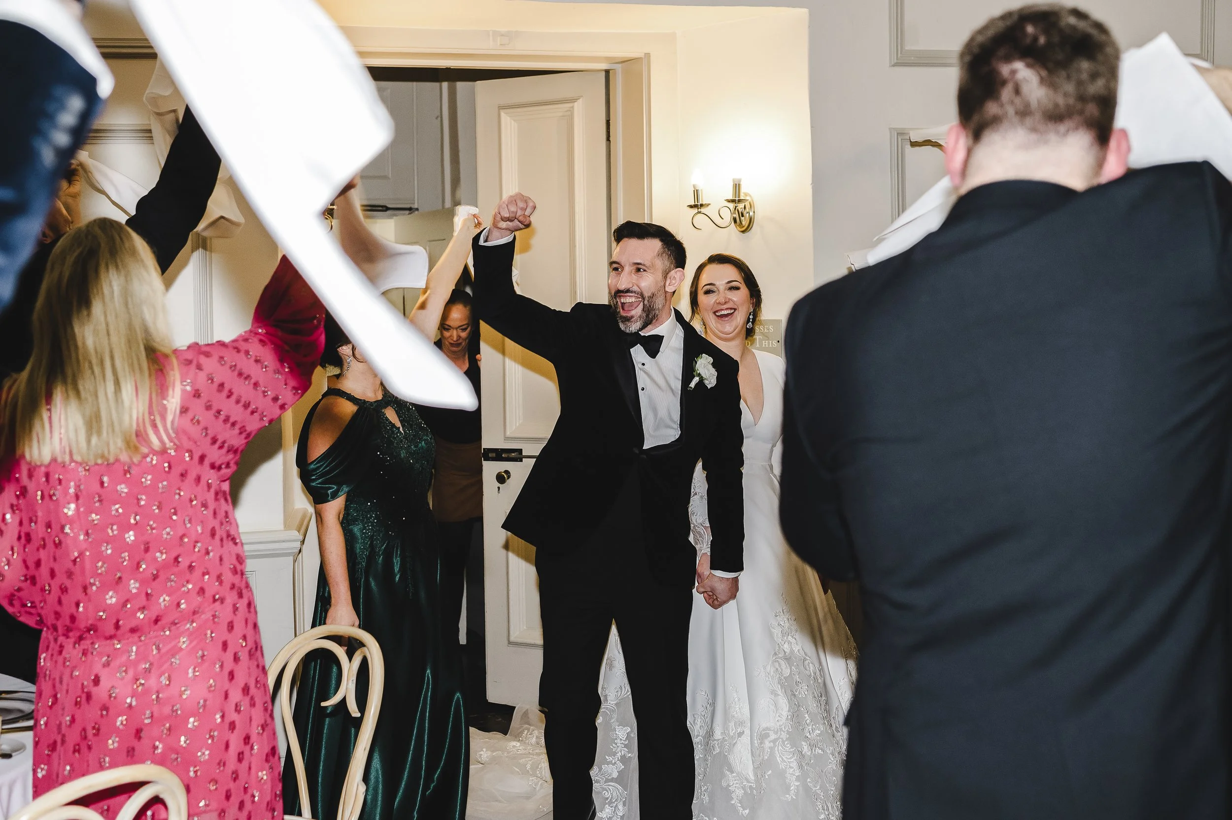 Bride & groom entering the wedding breakfast room, greeted by guests waving napkins