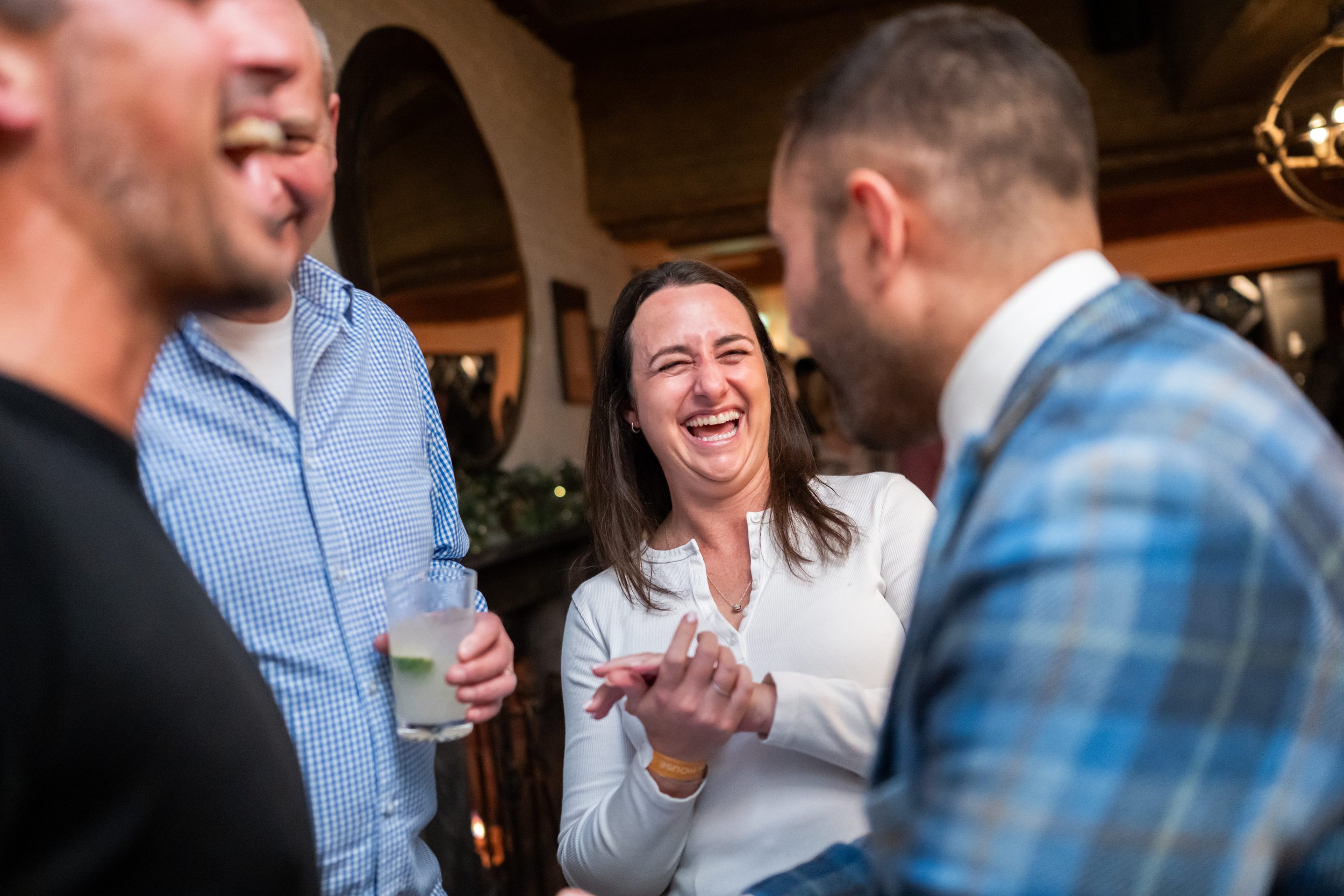 A professional magician in a tartan suit performs a close-up card trick for three smartly dressed people at a corporate drinks reception, one reacting with open-mouthed laughter