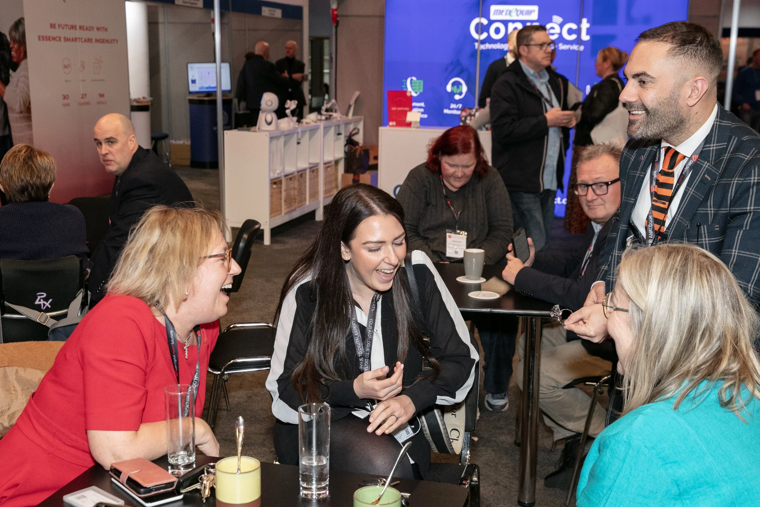 A magician performs close-up card magic for two female guests holding drinks at a busy networking event, drawing a look of astonished delight from one of the guests.