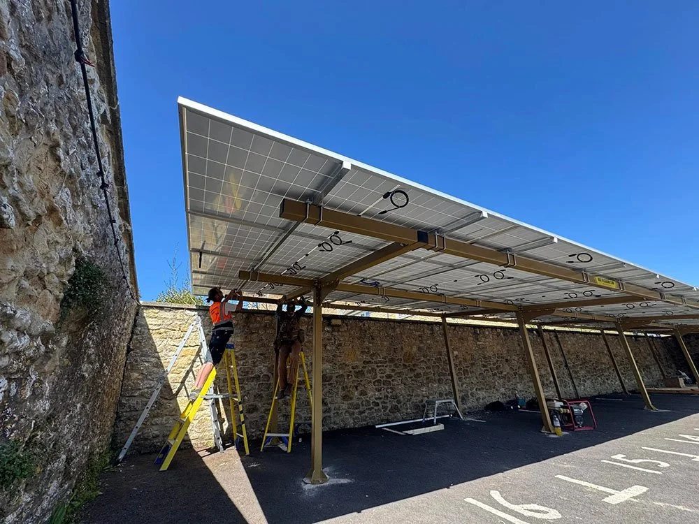 Two workers on ladders installing or maintaining solar panels on a metal structure in a parking lot, with a stone wall behind them and a clear blue sky above.