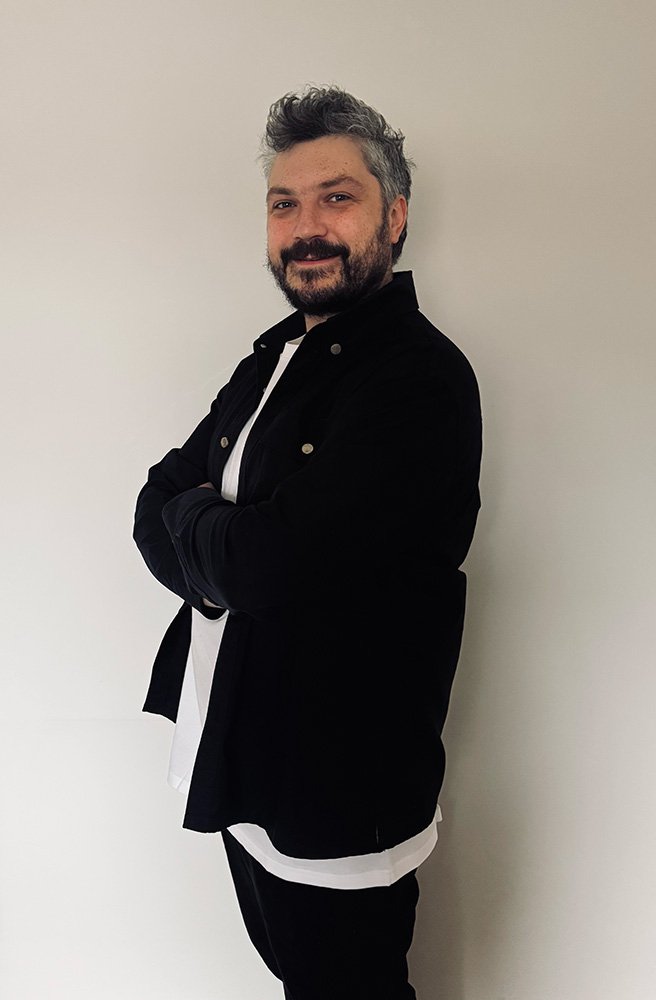 A man with gray hair and a beard, smiling with arms crossed, wearing a black jacket over a white shirt, standing against a plain white wall.