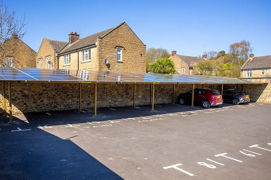 Parking lot with covered shade and several cars parked in front of a stone building with greenery.