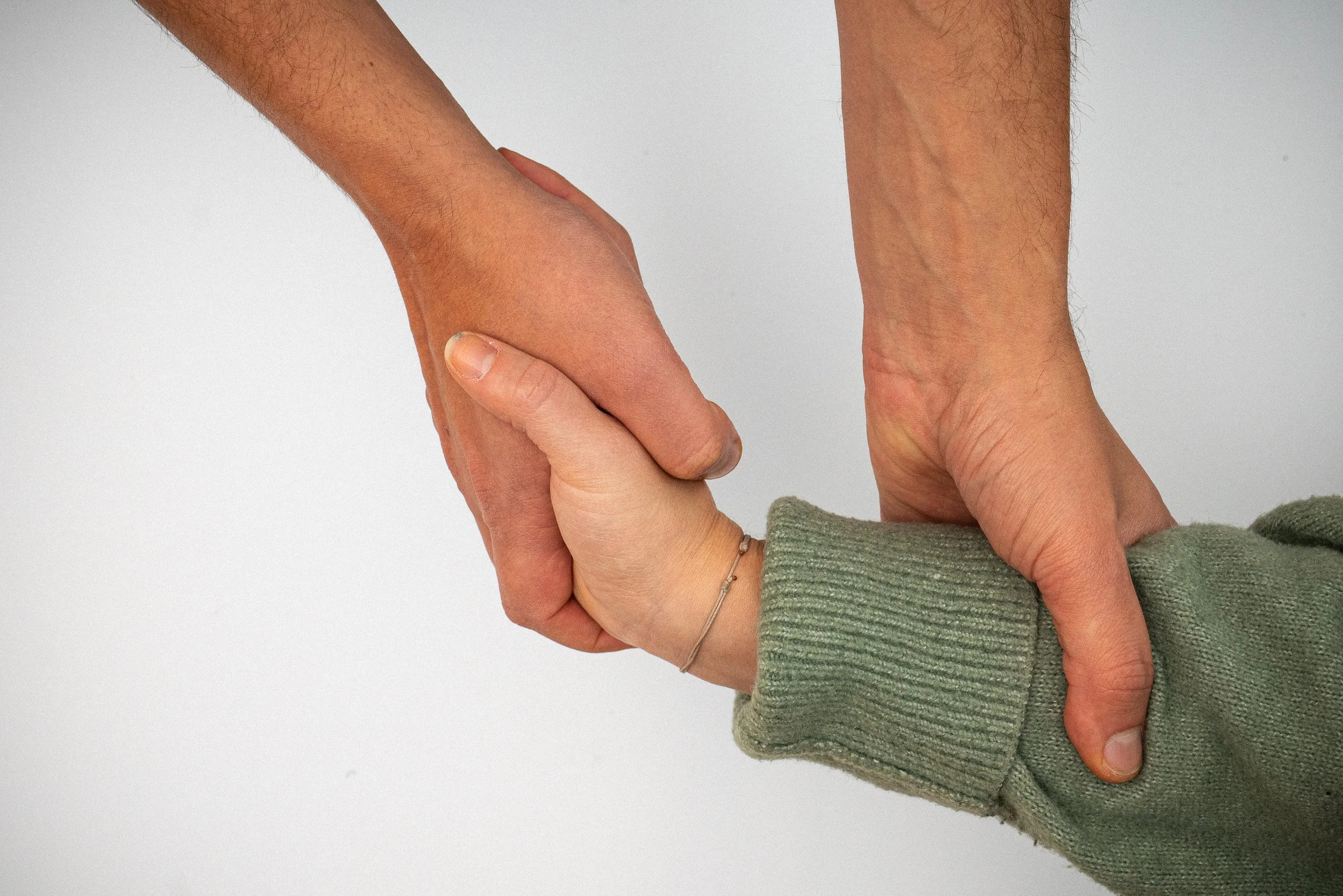 Zwei Menschen halten sich an der Hand, eine Person mit kürzeren Arm und heller Hautfarbe, die andere mit längeren Arm und etwas dunklerer Hautfarbe, gegen einen hellgrauen Hintergrund.