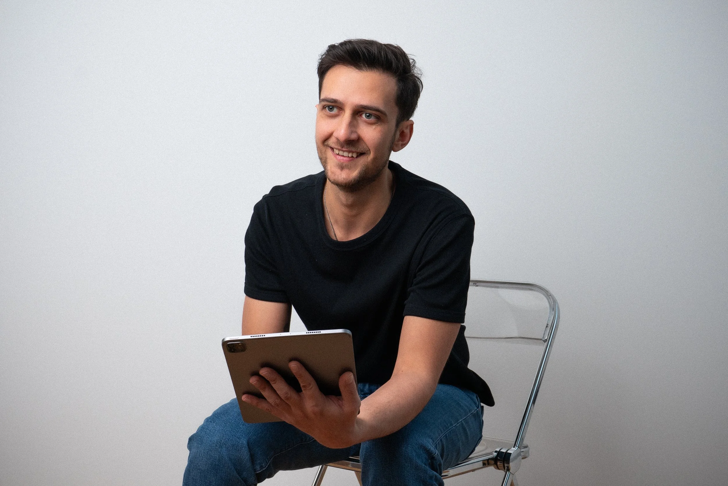 Junge Mann sitzt auf einem Stuhl vor einer weißen Wand und benutzt ein Tablet.