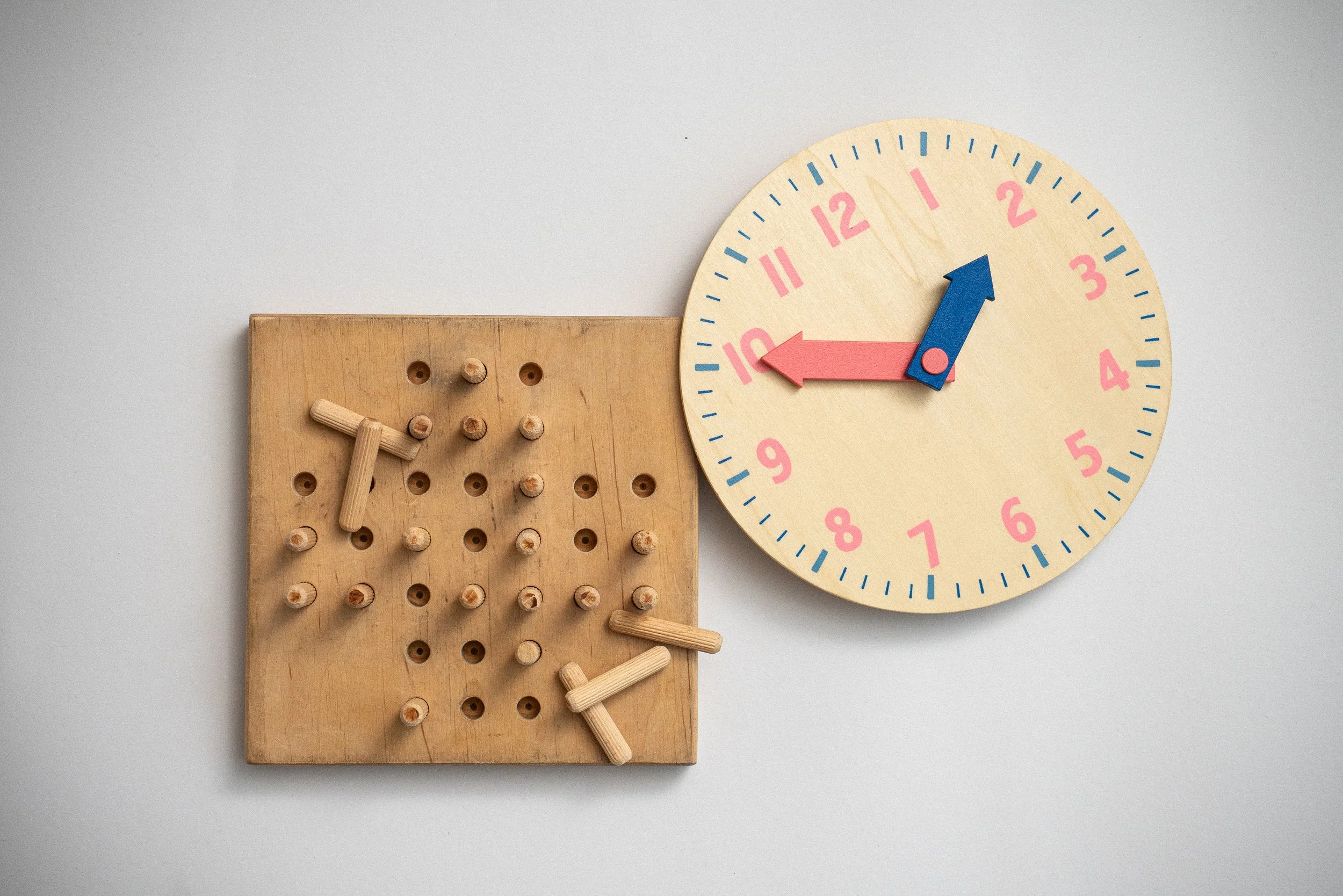 Ein Holzspielzeug-Uhr mit pinkem und blauer Uhrzeiger, daneben ein Holz-Puzzle mit Stäbchen.