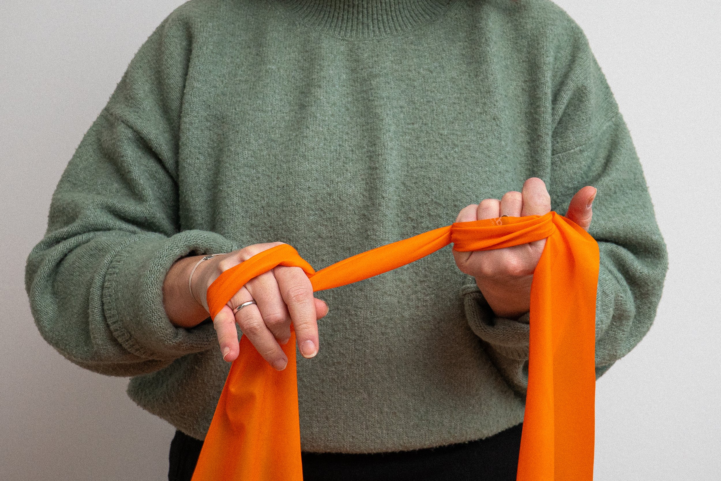 Person im grünen Pullover bindet orangefarbenes Stoffband zu einem Knoten.