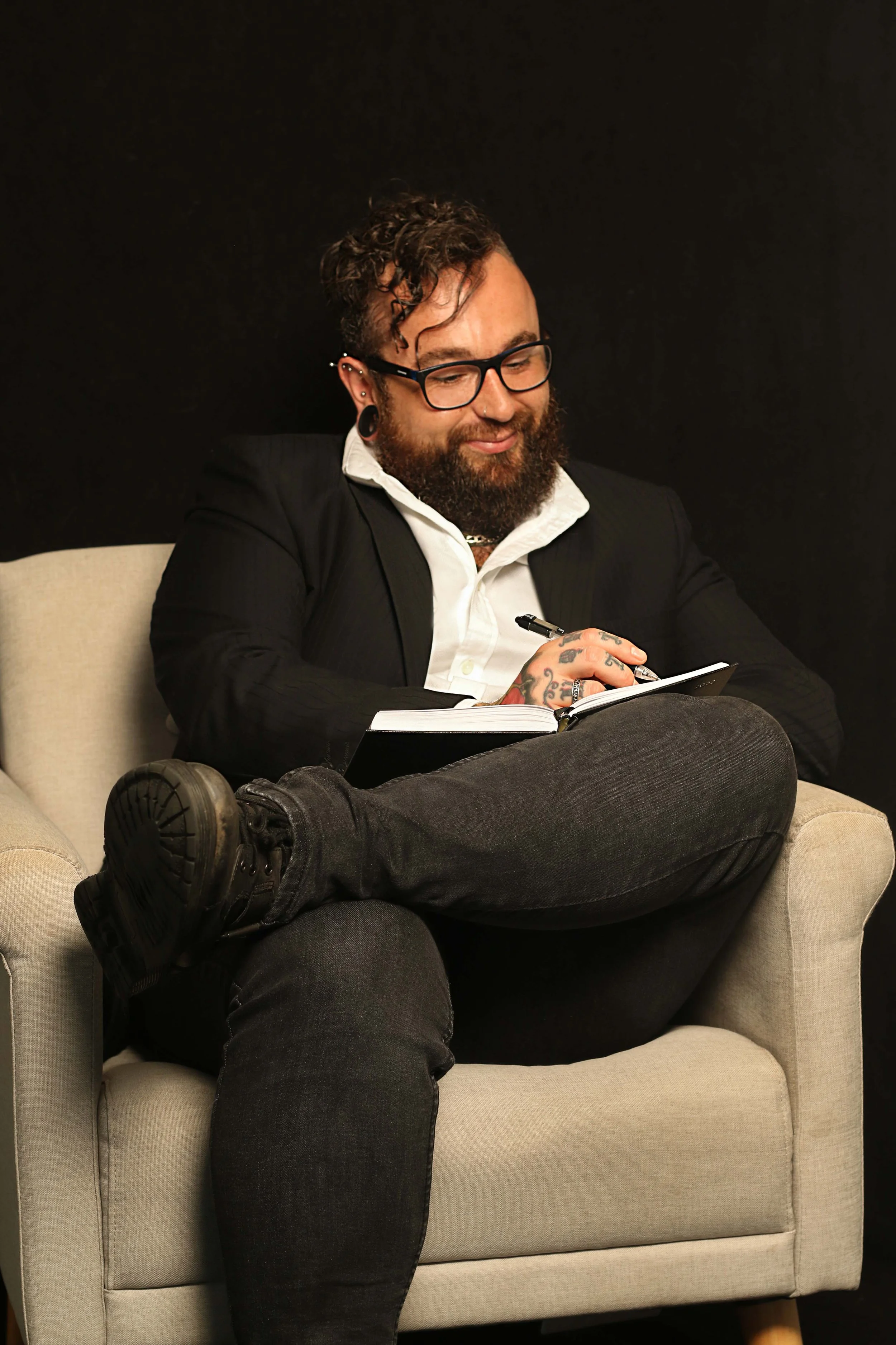 A man with glasses, tattoos, and earrings sits cross-legged on a beige armchair against a black background, writing in a notebook.