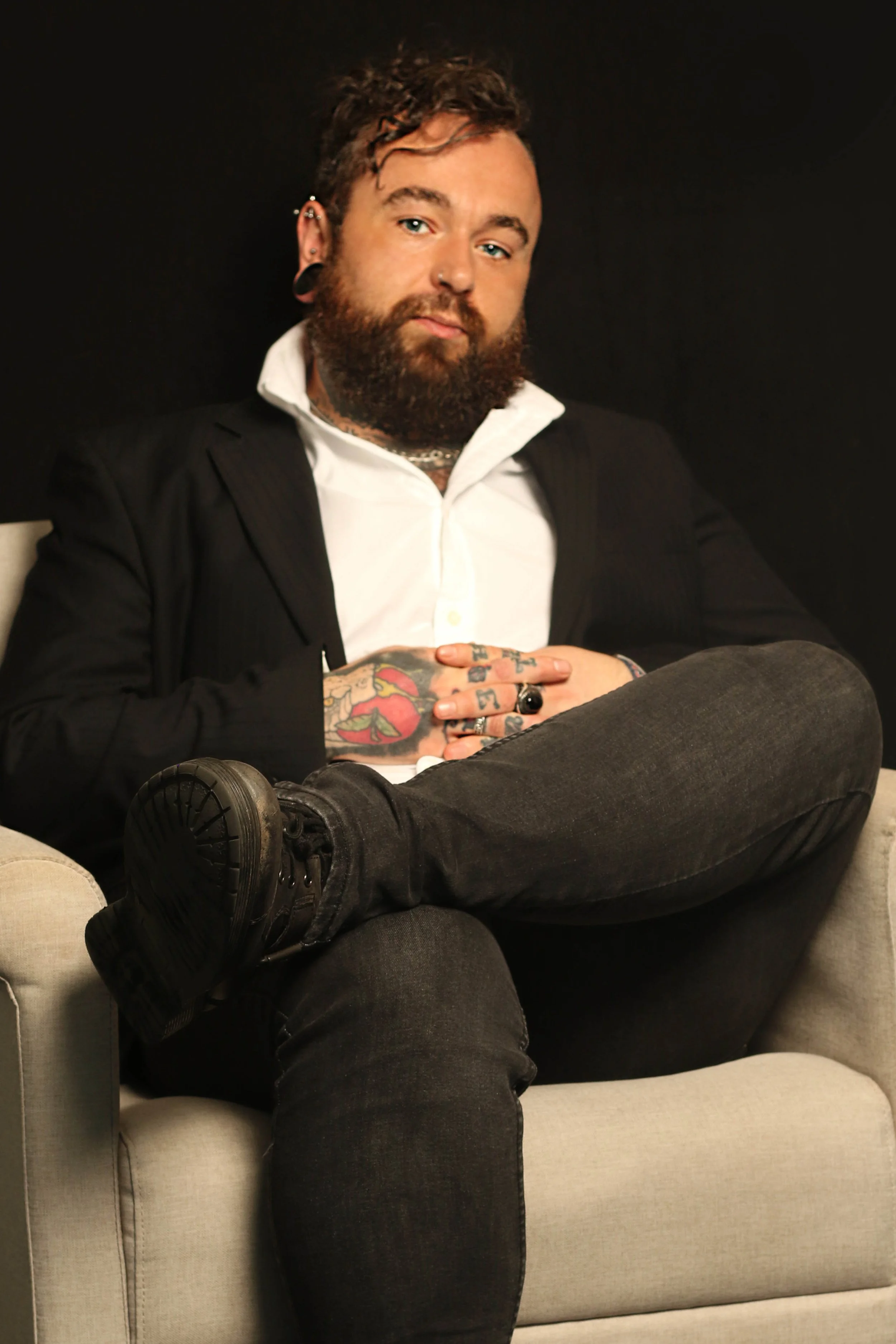 A man with a beard and tattoos sitting on a light-colored sofa, dressed in black jeans, a black blazer, and a white shirt, with his hands on his stomach and crossed legs, against a dark background.