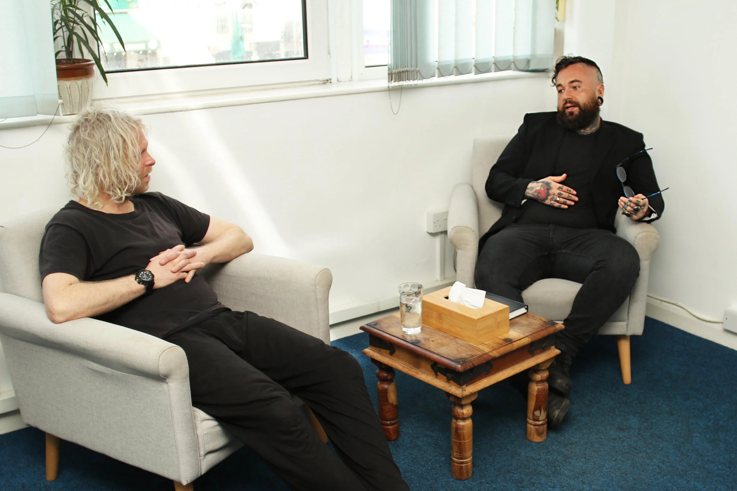 Two men sitting in a small office or lounge area. One with blonde curly hair and black t-shirt, the other with dark hair, beard, tattoos, and black clothing. They are having a conversation, seated on separate chairs with a wooden table between them, which has a glass of water and tissue box.